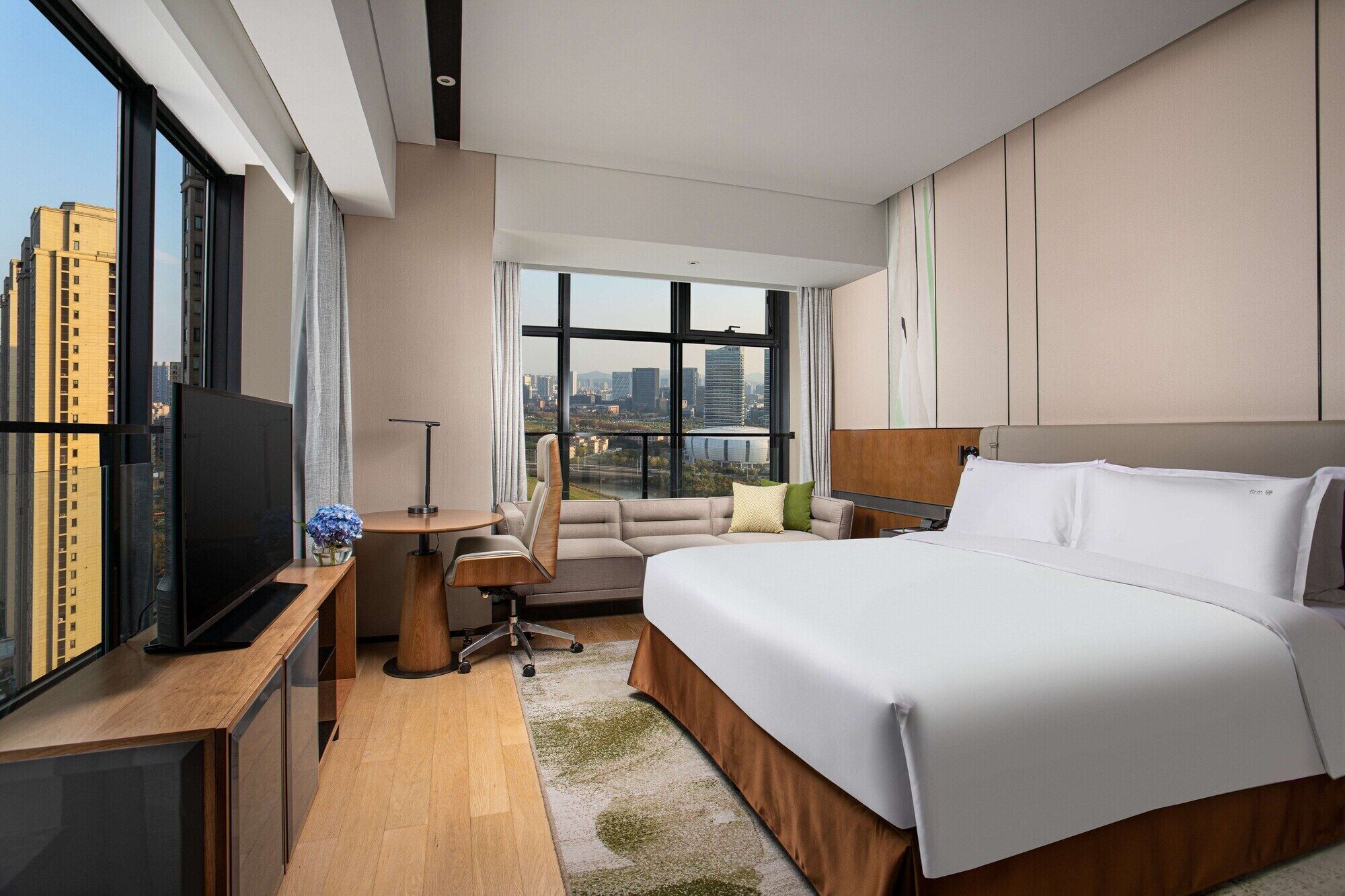 A modern hotel room with a large bed, a desk, a chair, a TV, and a view of the cityscape.