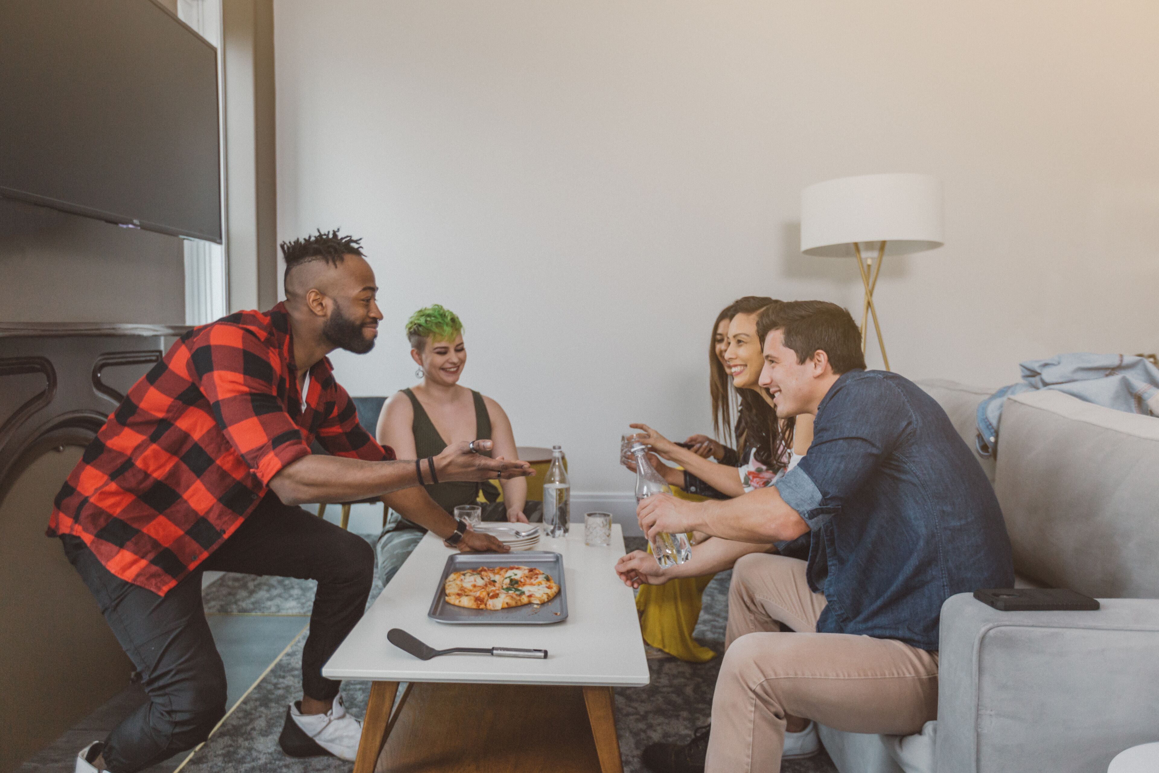 A group of people around a table with pizza, glasses, and bottles, in a room with a couch and a lamp.