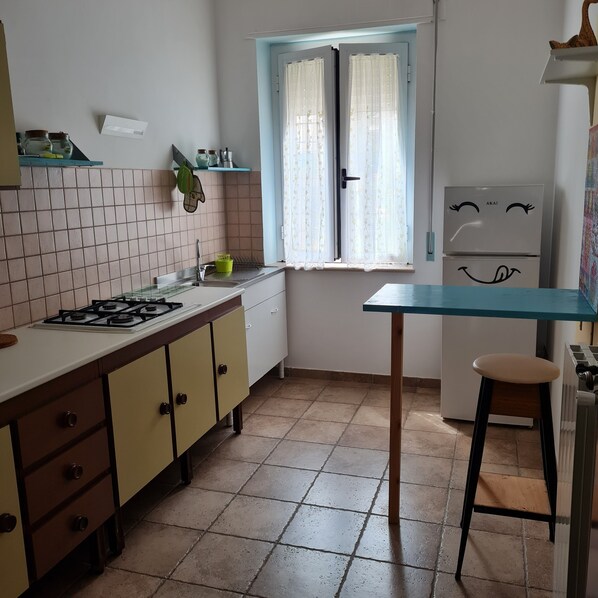 A kitchen with a gas stove, a sink, a window with curtains, a refrigerator, and a stool.