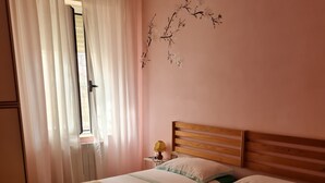 Quarto de hotel com duas camas, uma janela com cortinas leves, um adorno de galho fixado na parede e um abajur.