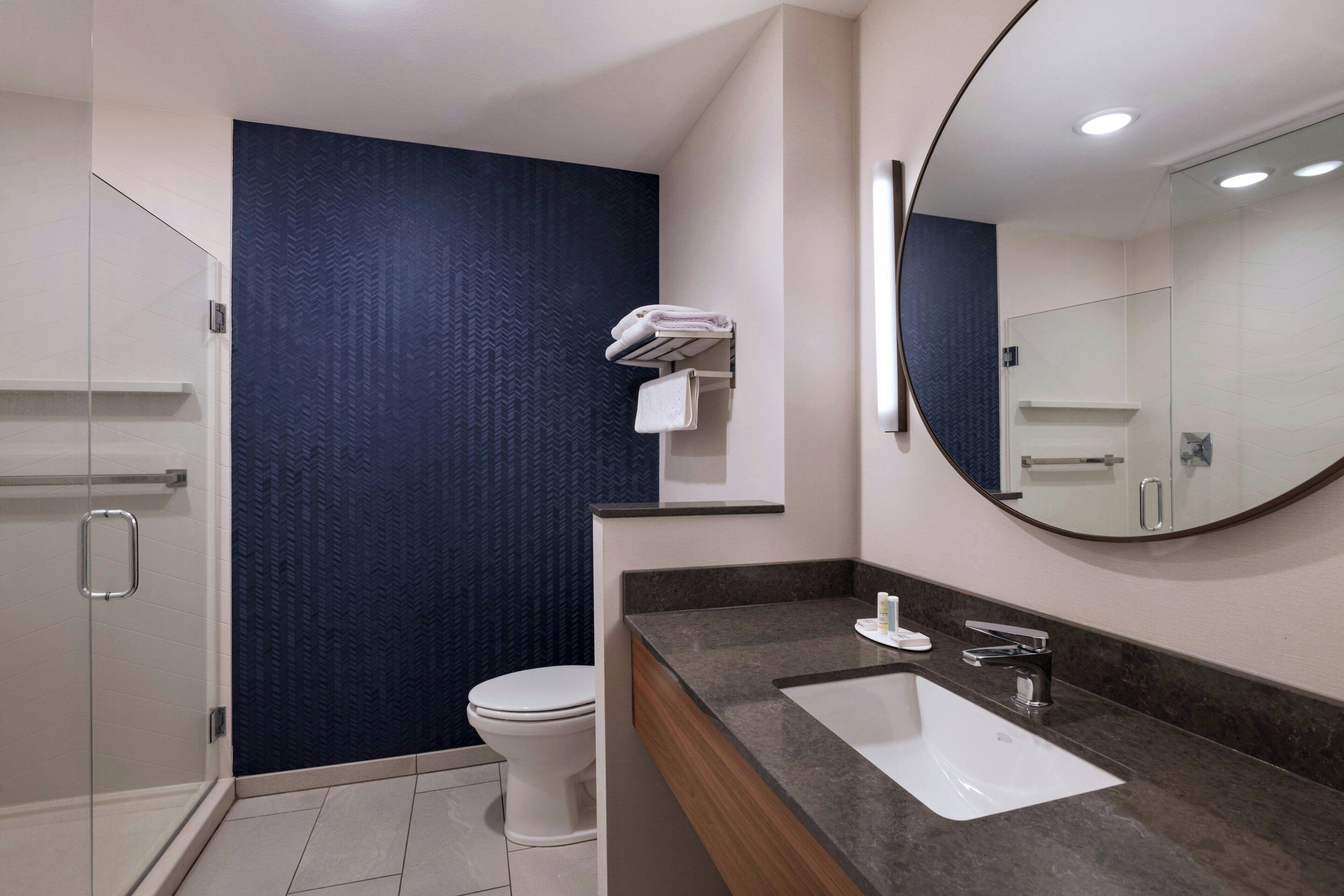 Een moderne badkamer met een glazen douchecabine, een wit toilet, een donkerblauwe wand, een witte wastafel met een marmeren aanrechtblad en een grote spiegel.