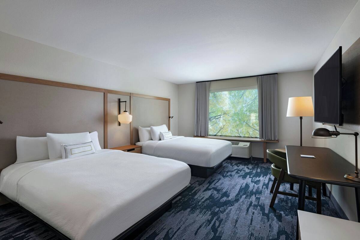 A hotel room with two beds, a desk, a chair, a TV, and a window with curtains.