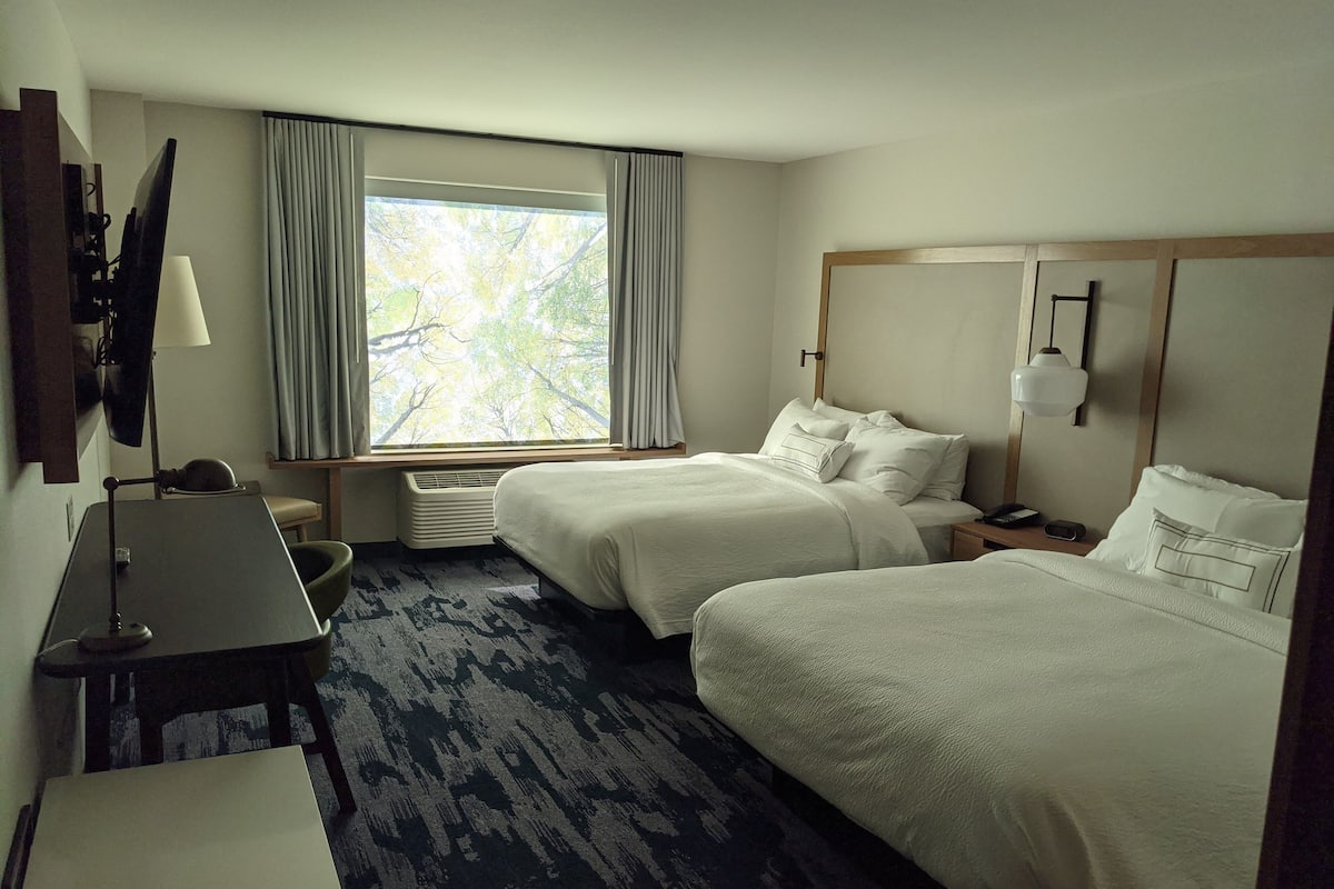 A hotel room with two beds, a desk, a chair, a lamp, a window with curtains, and a view of trees.