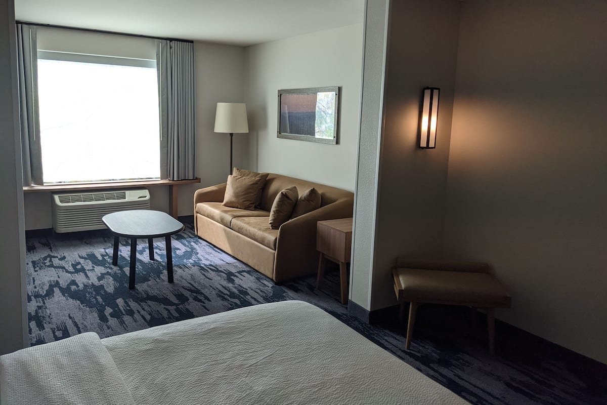 A hotel room with a bed, a sofa, a small table, a lamp, and a window with curtains.