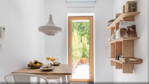 A dining area with a wooden table, chairs, a wicker pendant light, and a view of greenery through a door.