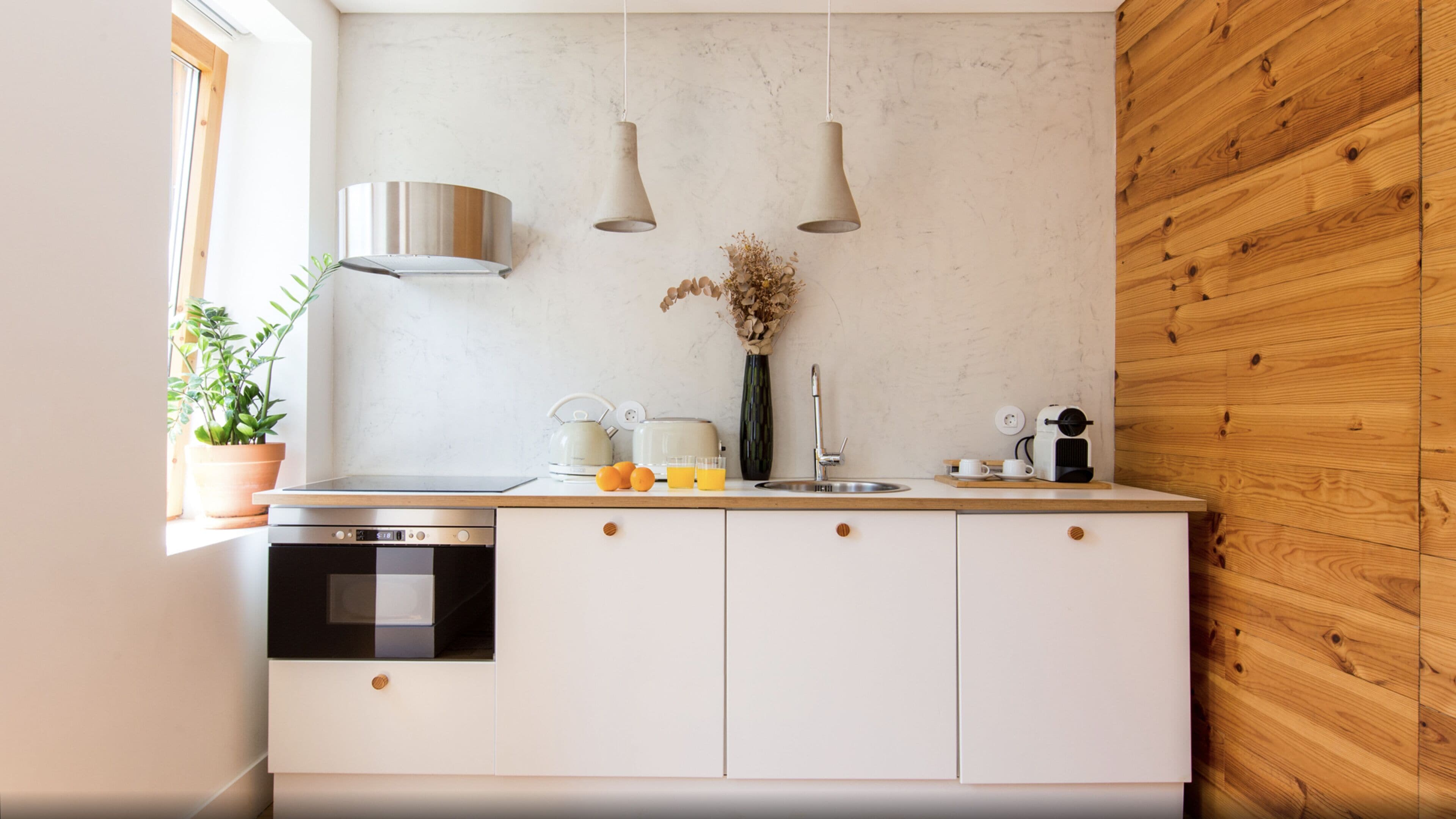 A modern kitchen with white cabinets, a stainless steel range hood, a built-in oven, a sink, and a wooden wall.