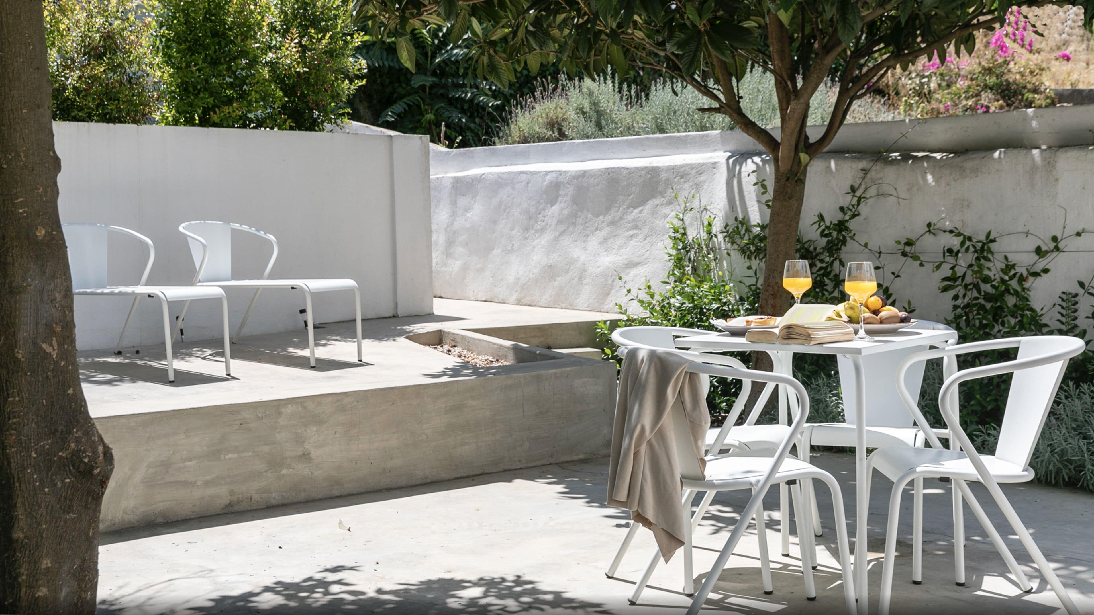 Un patio exterior con sillas y una mesa blancas dispuestas para una comida, rodeado de follaje y una pared blanca.