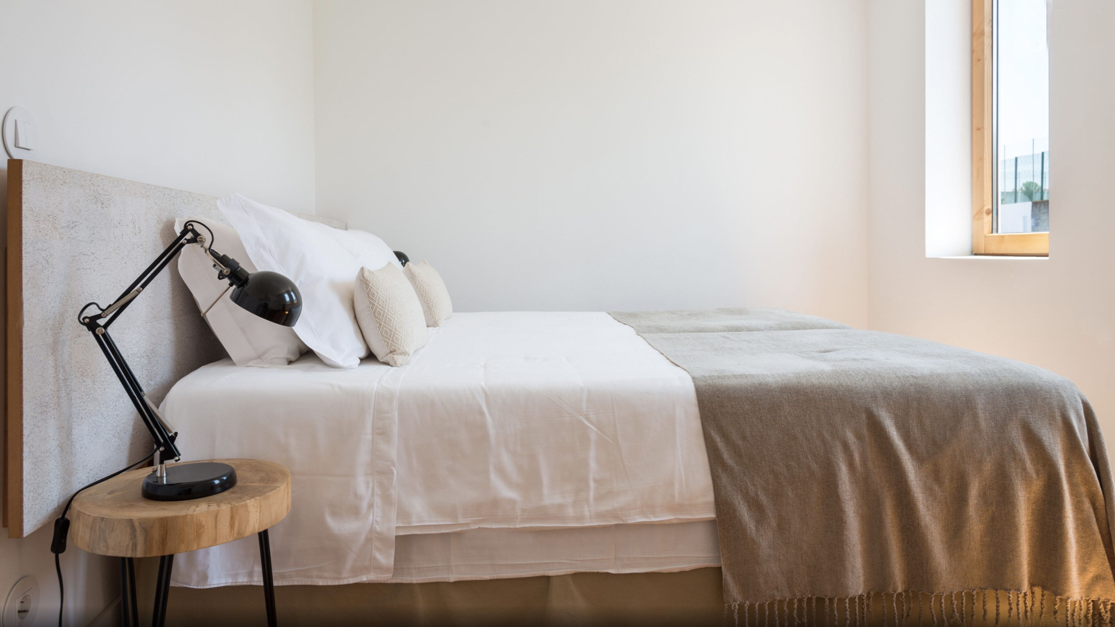 A neatly made bed with white bedding and a beige throw blanket, a small round wooden nightstand with a lamp, and a window with a view of a building.