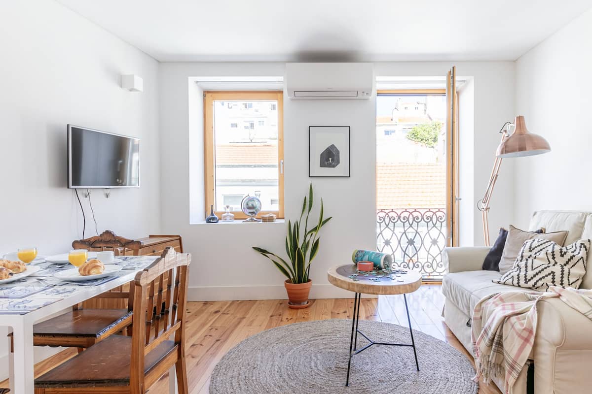 Une pièce bien éclairée, avec une table à manger dressée pour le petit-déjeuner, un canapé avec une couverture, un téléviseur fixé au mur et une grande fenêtre donnant sur des immeubles.