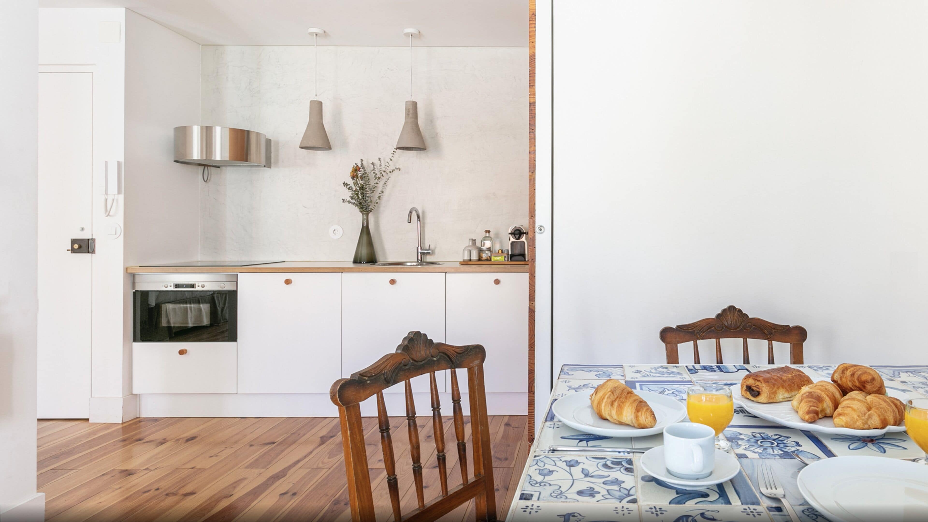 Eine moderne Küche mit weißen Schränken, einem eingebauten Backofen und einer Edelstahl-Dunstabzugshaube. Es gibt einen Esstisch mit einer blau-weißen Tischdecke, einen Holzstuhl und einen Teller mit Croissants und Orangensaft.
