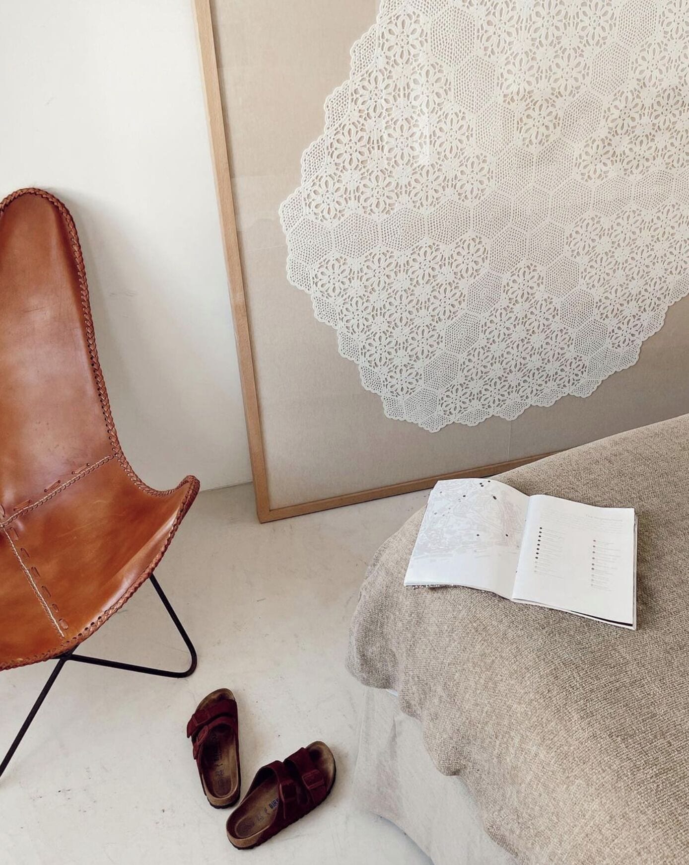 Quarto com uma poltrona de couro marrom, um par de chinelos marrons, um livro branco e uma obra de arte em renda branca emoldurada.