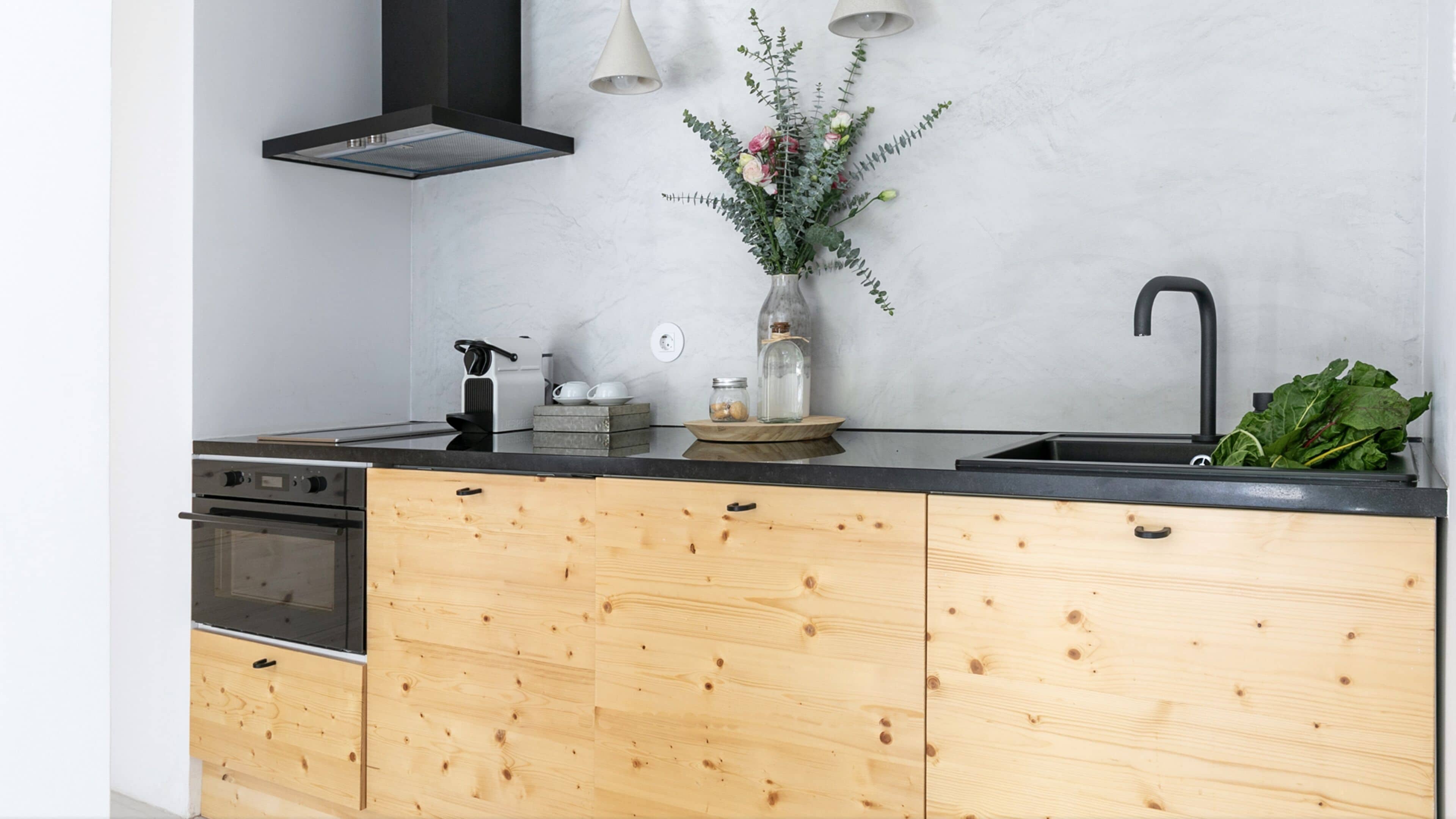 A modern kitchen with wooden cabinets, a black countertop, a built-in oven, a stainless steel range hood, a vase with flowers, and a sink with a faucet.