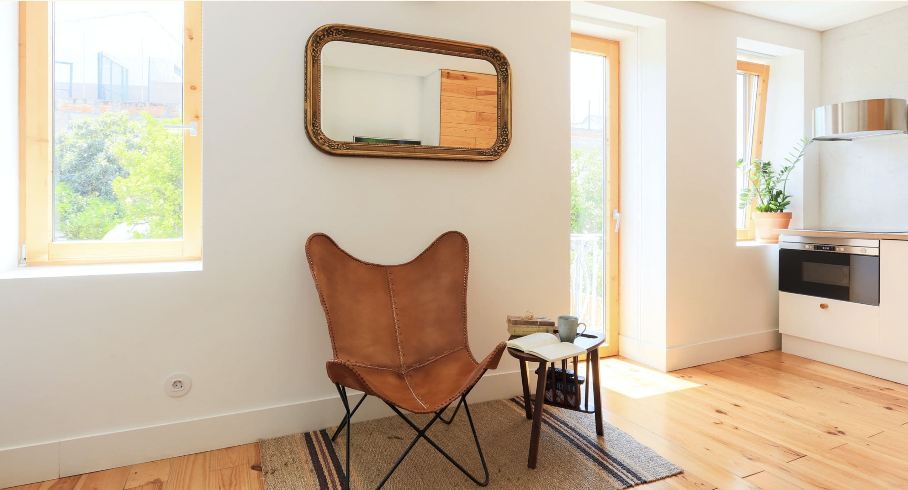 A room with a brown leather chair, a small table with a cup and a book, a mirror on the wall, and a kitchen area with a refrigerator and stove.