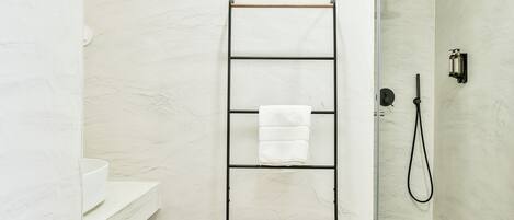 Een badkamer met een witte marmeren wand, een zwarte handdoekhouder met opgevouwen handdoeken, een douche met een handdouche en een wit wastafelmeubel.