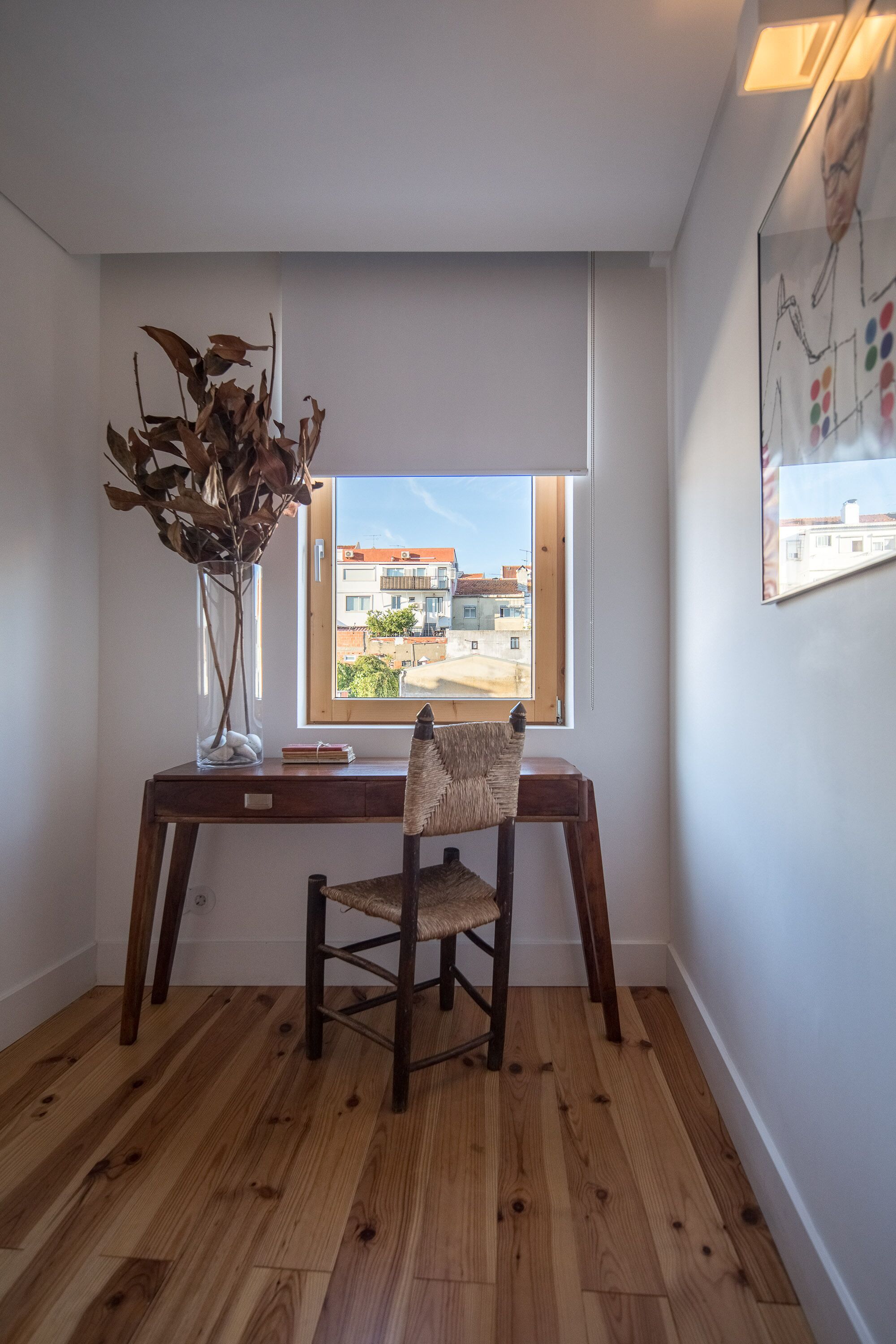 A room with a wooden desk, a chair, a vase with dried leaves, and a window with a view of buildings.