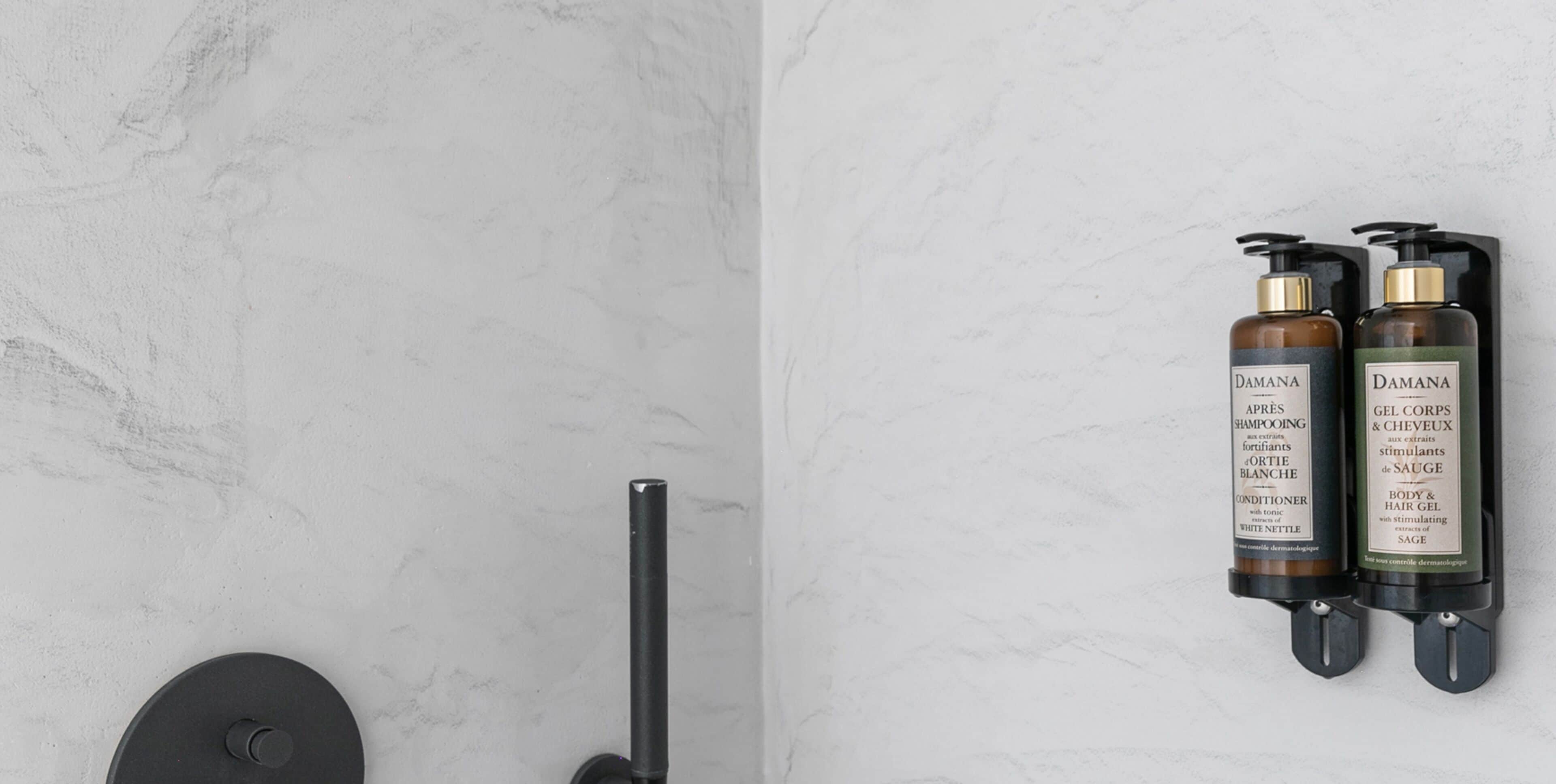 Casa de banho com parede de mármore, torneira preta e dois frascos de produtos Damana para corpo e cabelo.