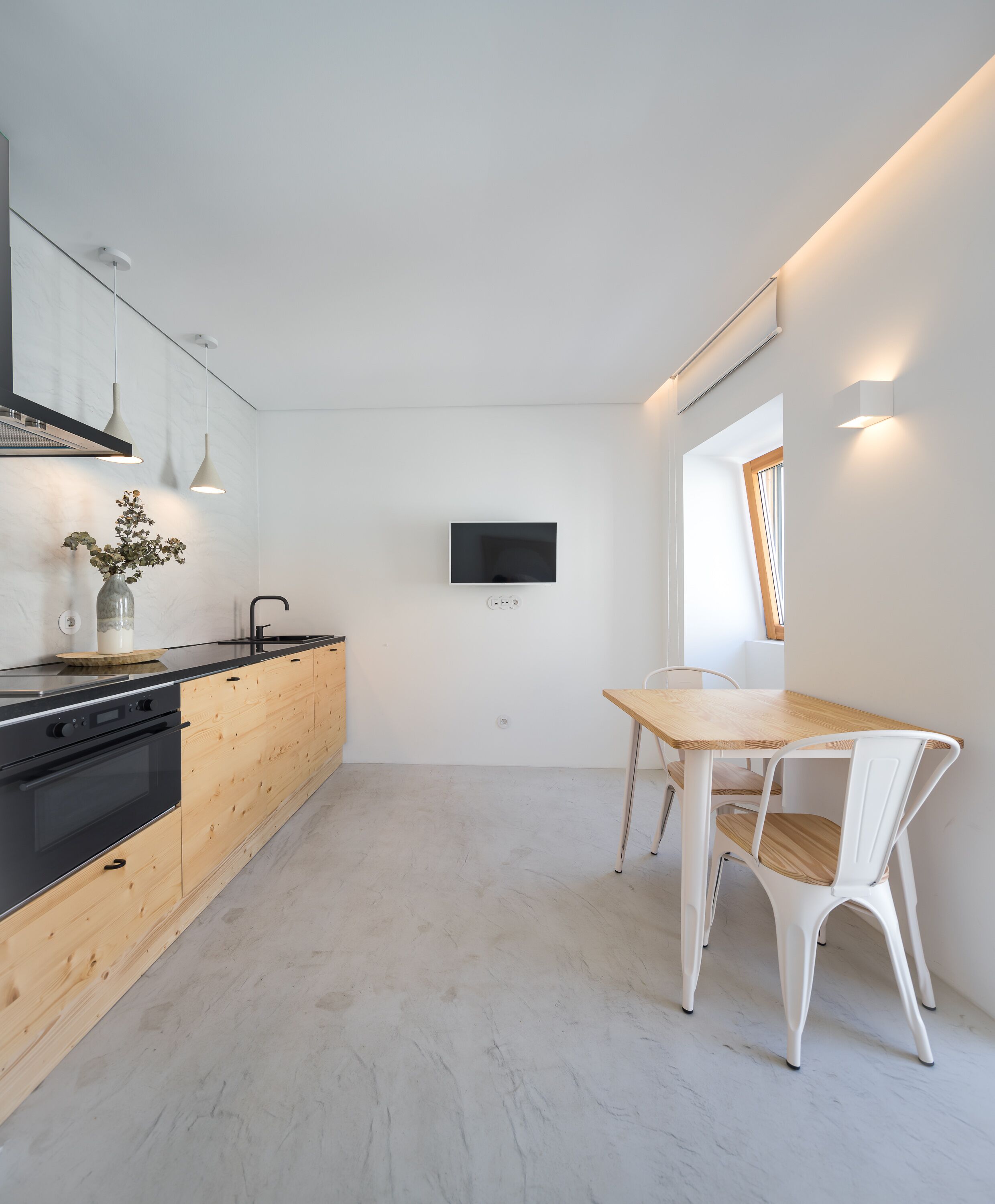 Una cocina moderna con armarios de madera, estufa negra, luces colgantes, televisor montado en la pared y un comedor con mesa de madera y sillas blancas.