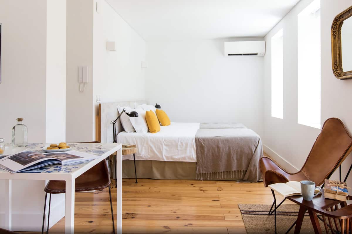Une chambre à coucher moderne et minimaliste, avec un plancher en bois, un lit agrémenté d’oreillers blancs et jaunes, un bureau avec une chaise et un grand miroir.