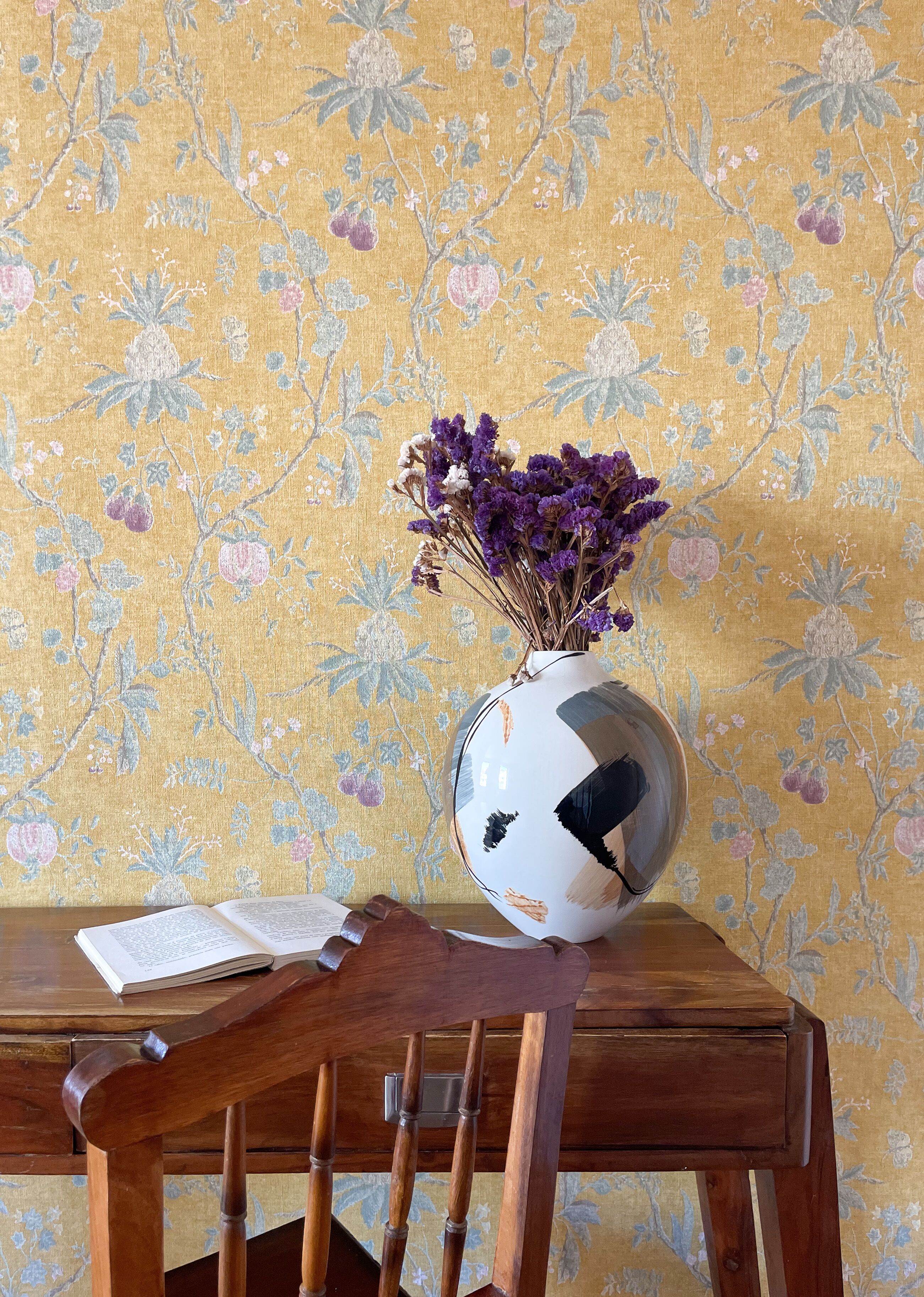 A wooden table with a white vase containing purple flowers, a book, and a wooden chair.