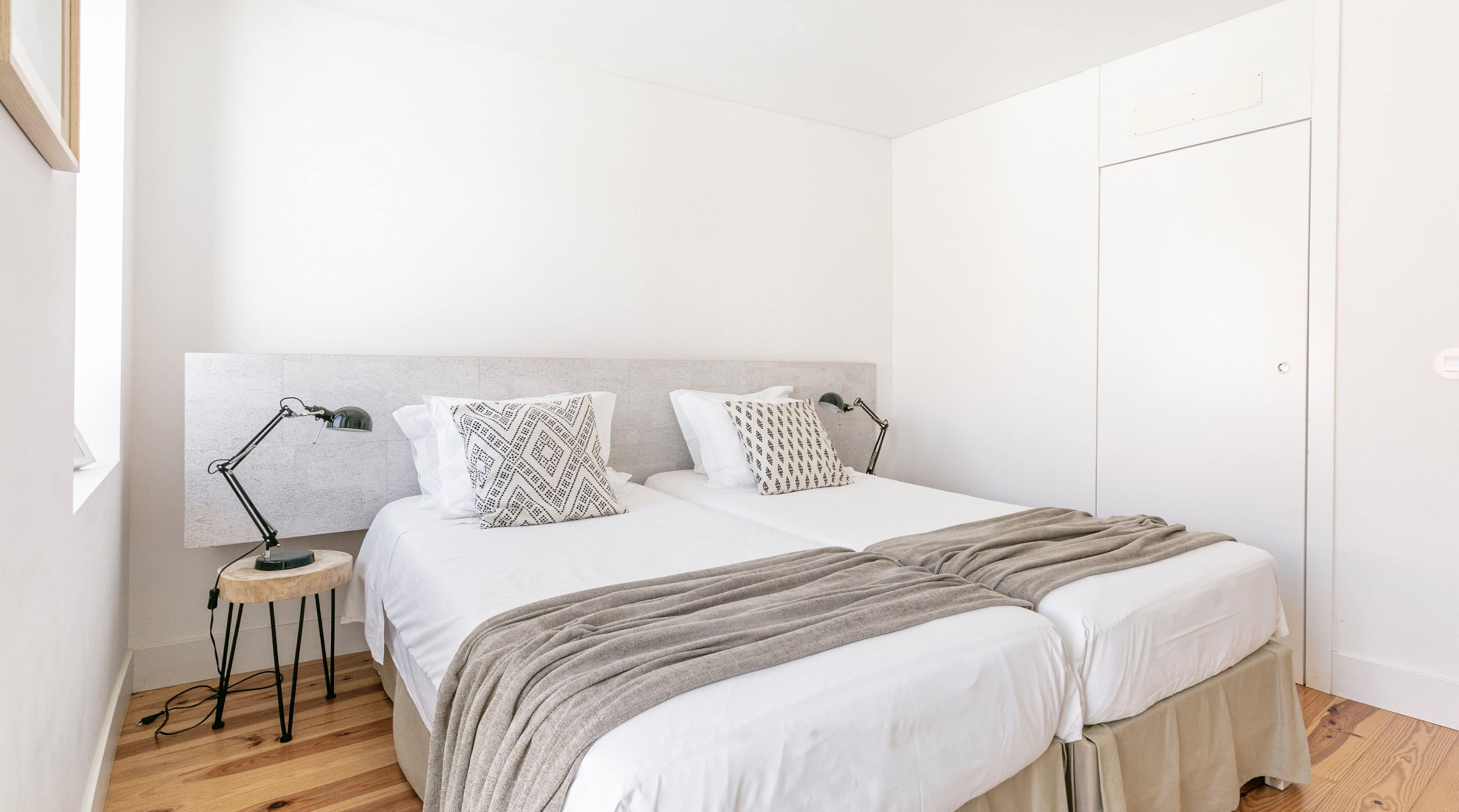 Un lit double avec du linge de lit blanc, deux coussins à motifs, un plaid gris et une table de chevet avec une lampe.