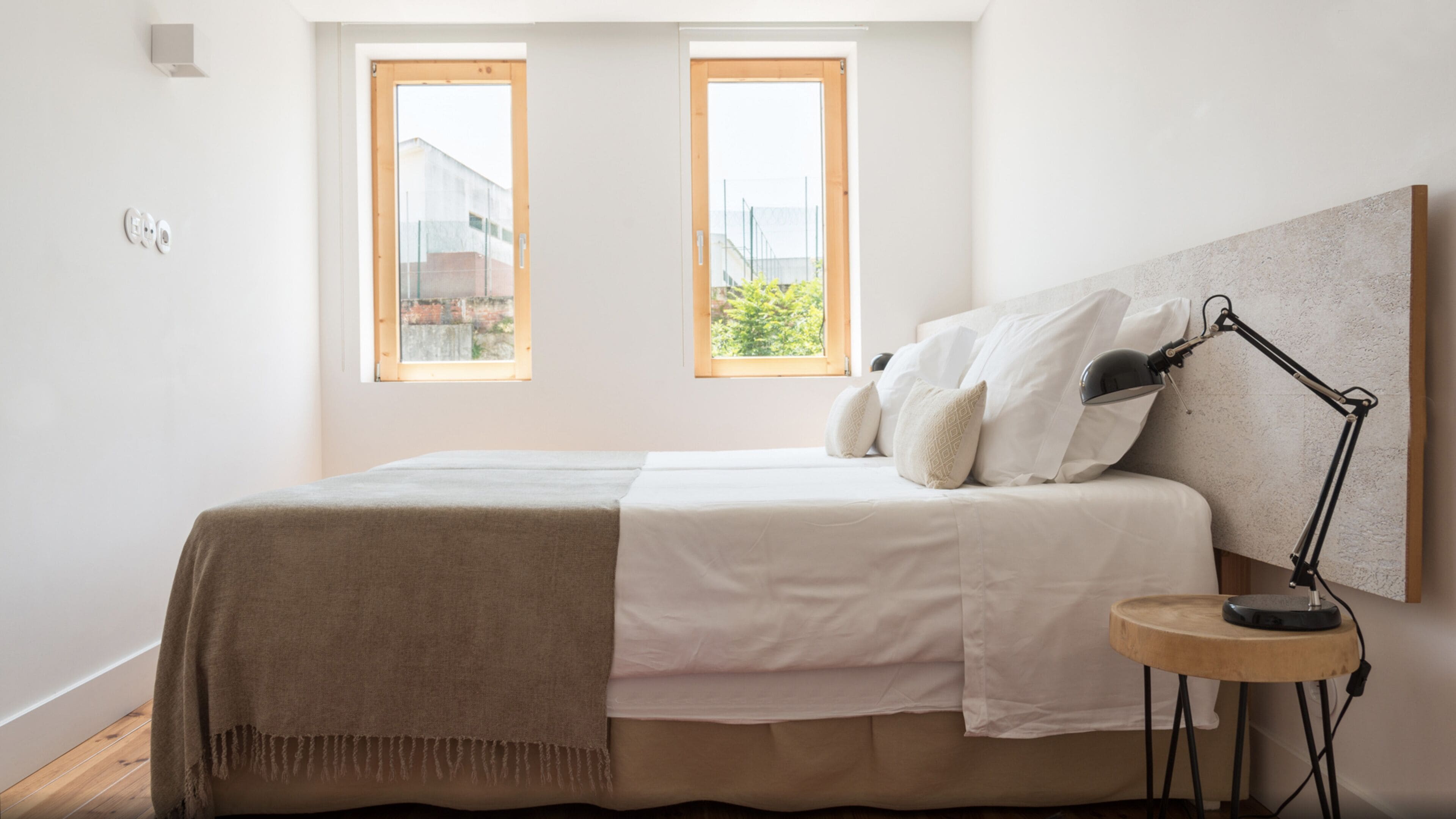 Una camera da letto con un letto, due finestre, un comodino con una lampada e un punto luce a parete.