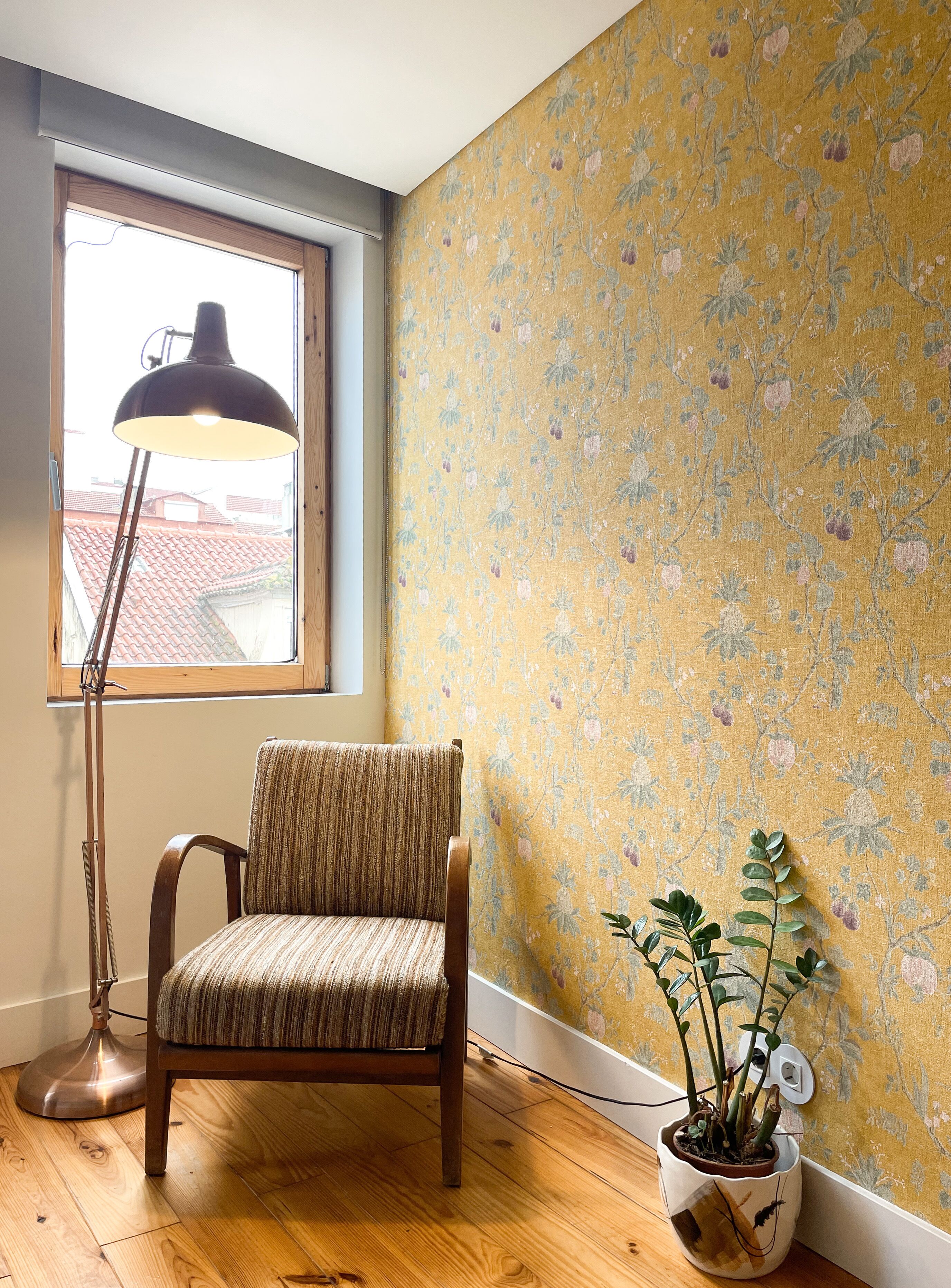 A corner room with a wooden floor, a chair with a striped cushion, a floor lamp, a potted plant, and a floral wallpaper.