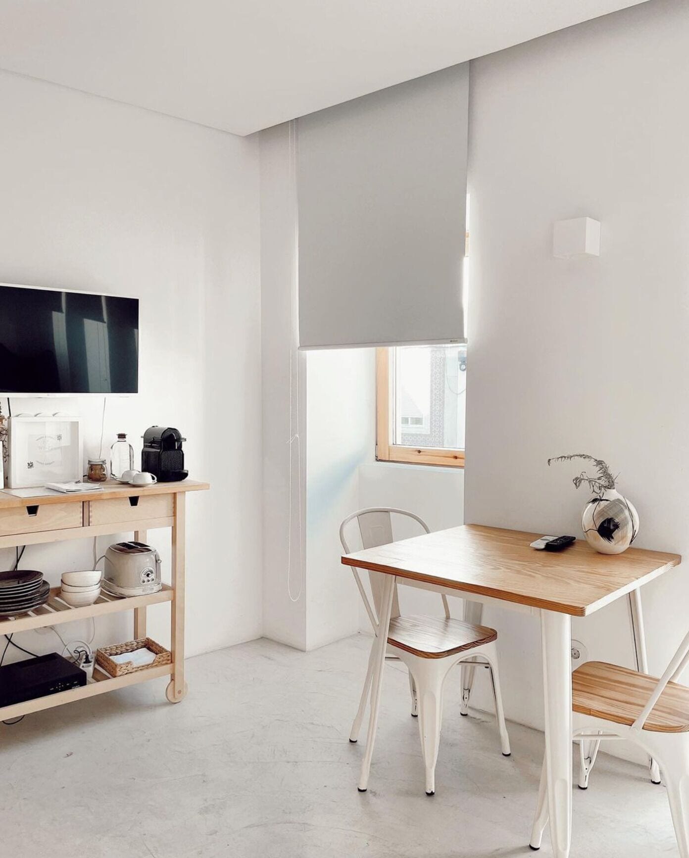 A minimalist room with a wooden desk, white chairs, a flat-screen TV, and a window with a view.