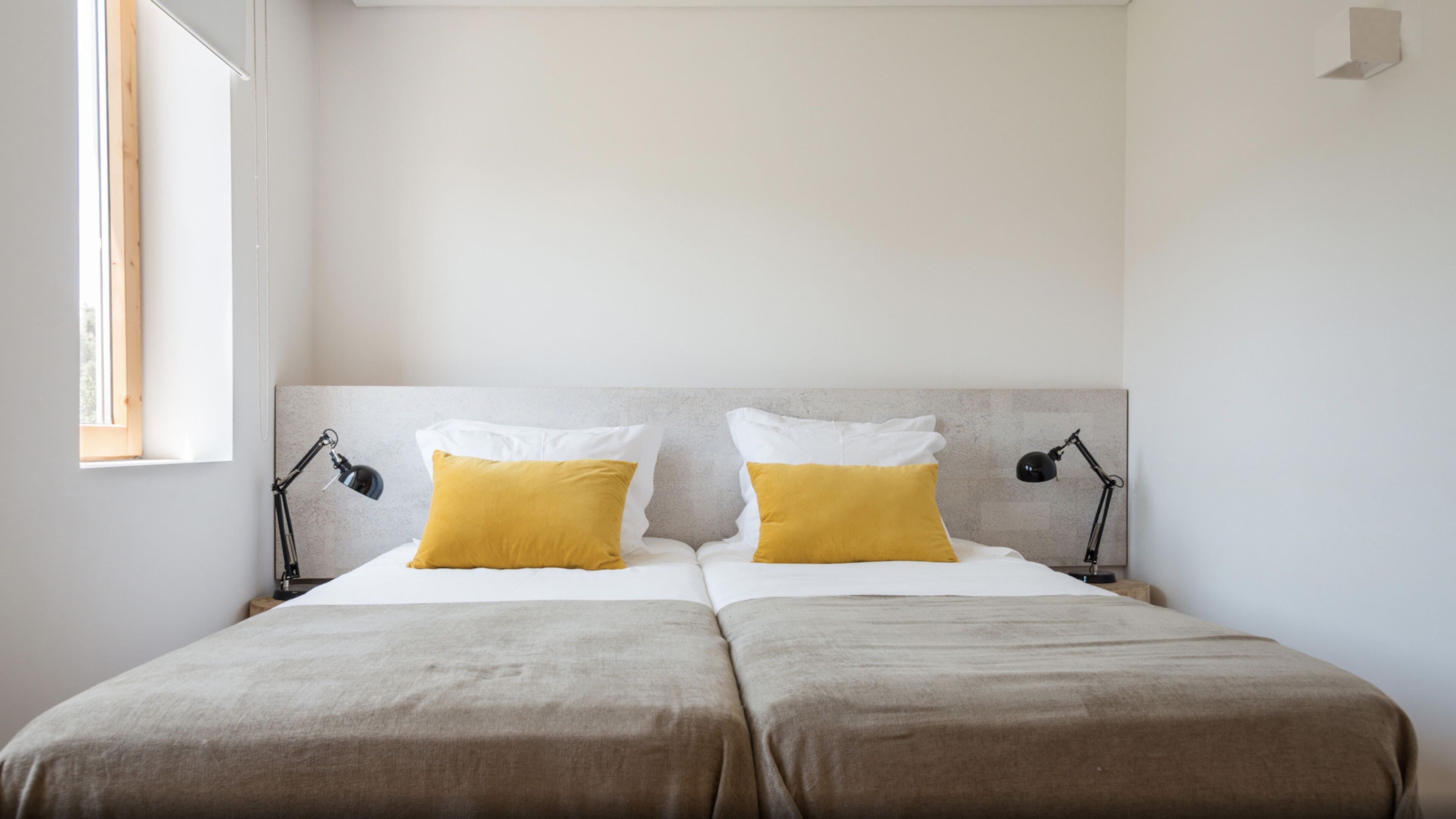 Un lit double avec deux oreillers blancs et deux oreillers de couleur jaune moutarde, flanqué de deux lampes de bureau noires.