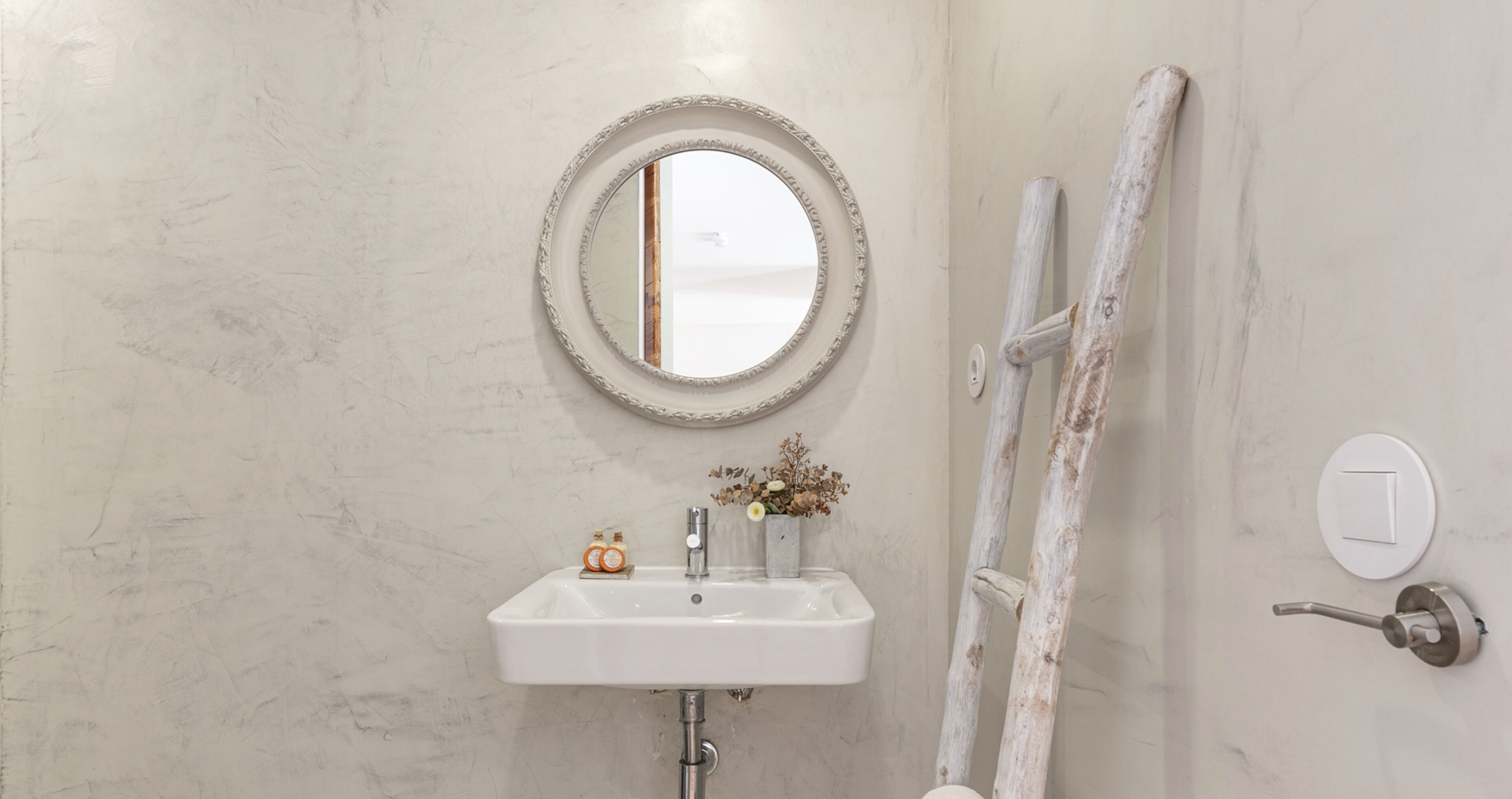 A bathroom with a white sink, a round mirror, and a wooden ladder leaning against the wall.