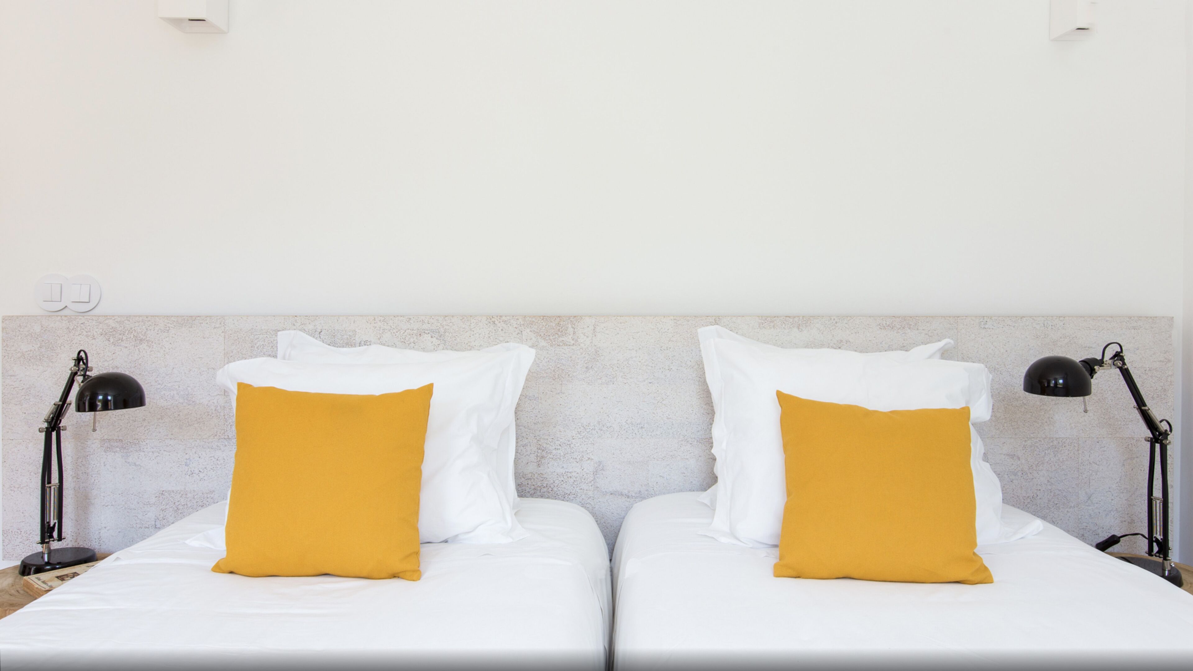 A double bed with white bedding and two mustard-colored pillows, flanked by black desk lamps on either side.