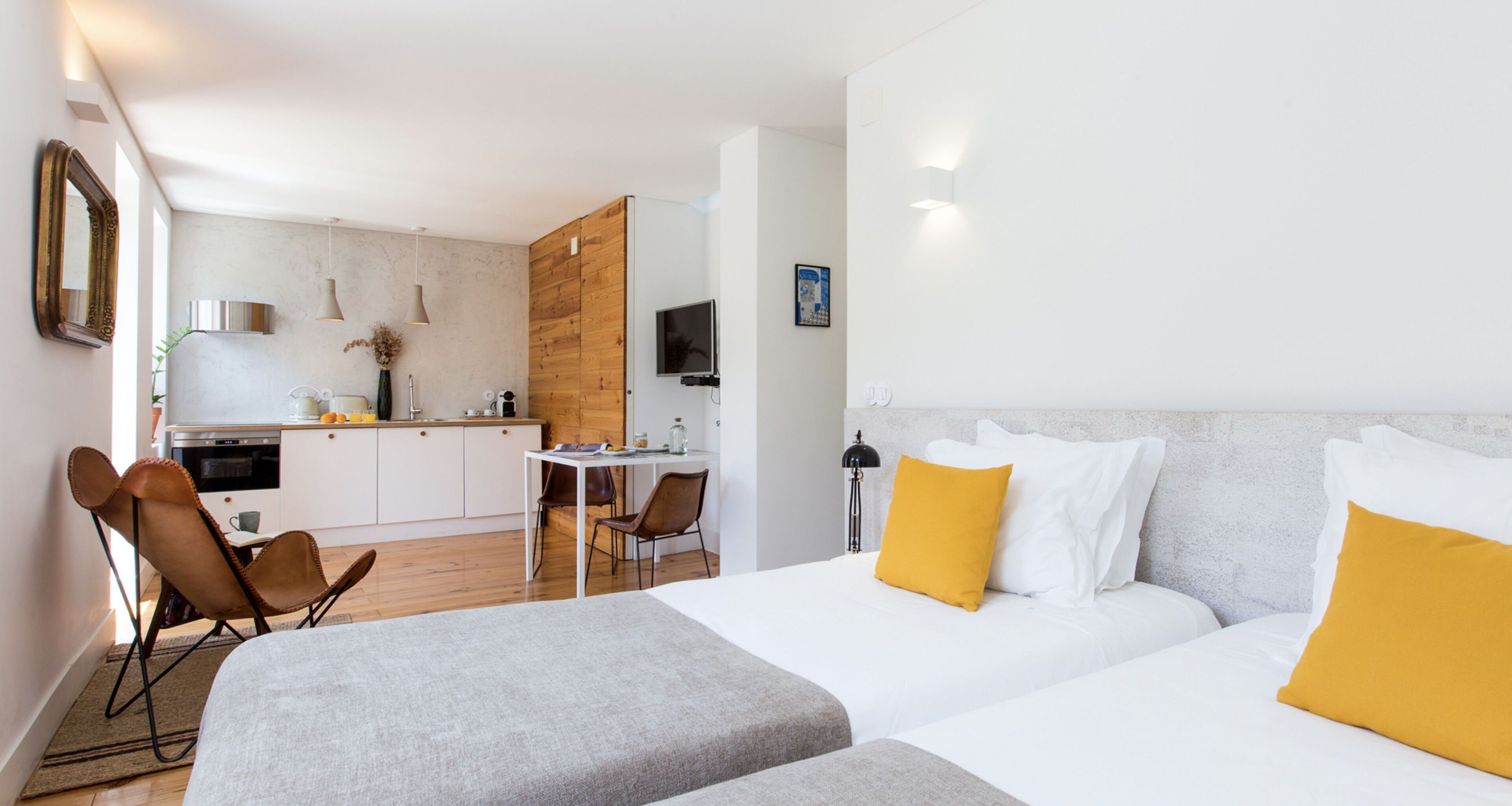 A modern hotel room with a large bed, a small desk, and a kitchenette in the background.