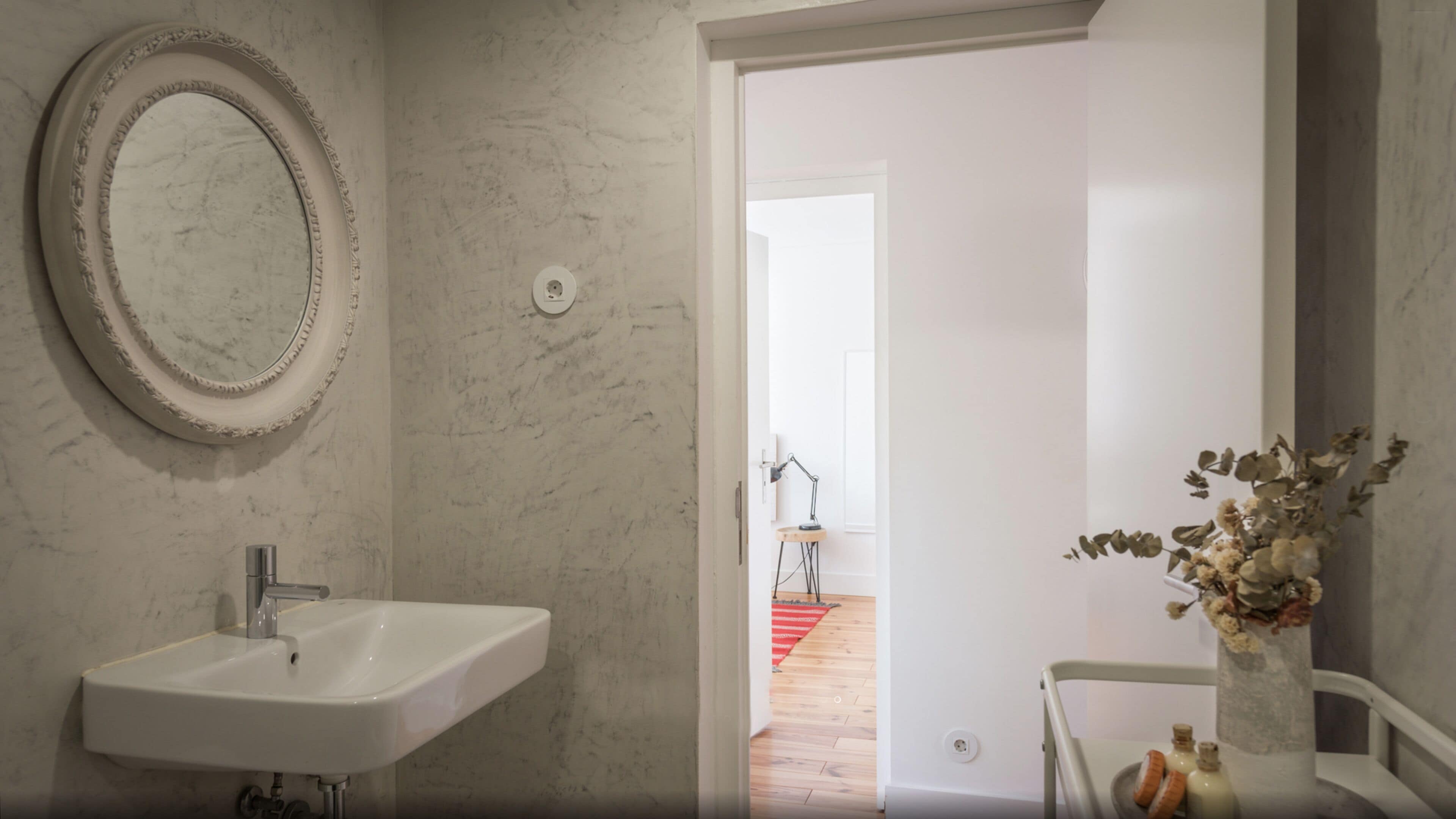 Ein Badezimmer mit einem weißen Waschbecken, einem runden Spiegel und einer Vase mit getrockneten Blumen.