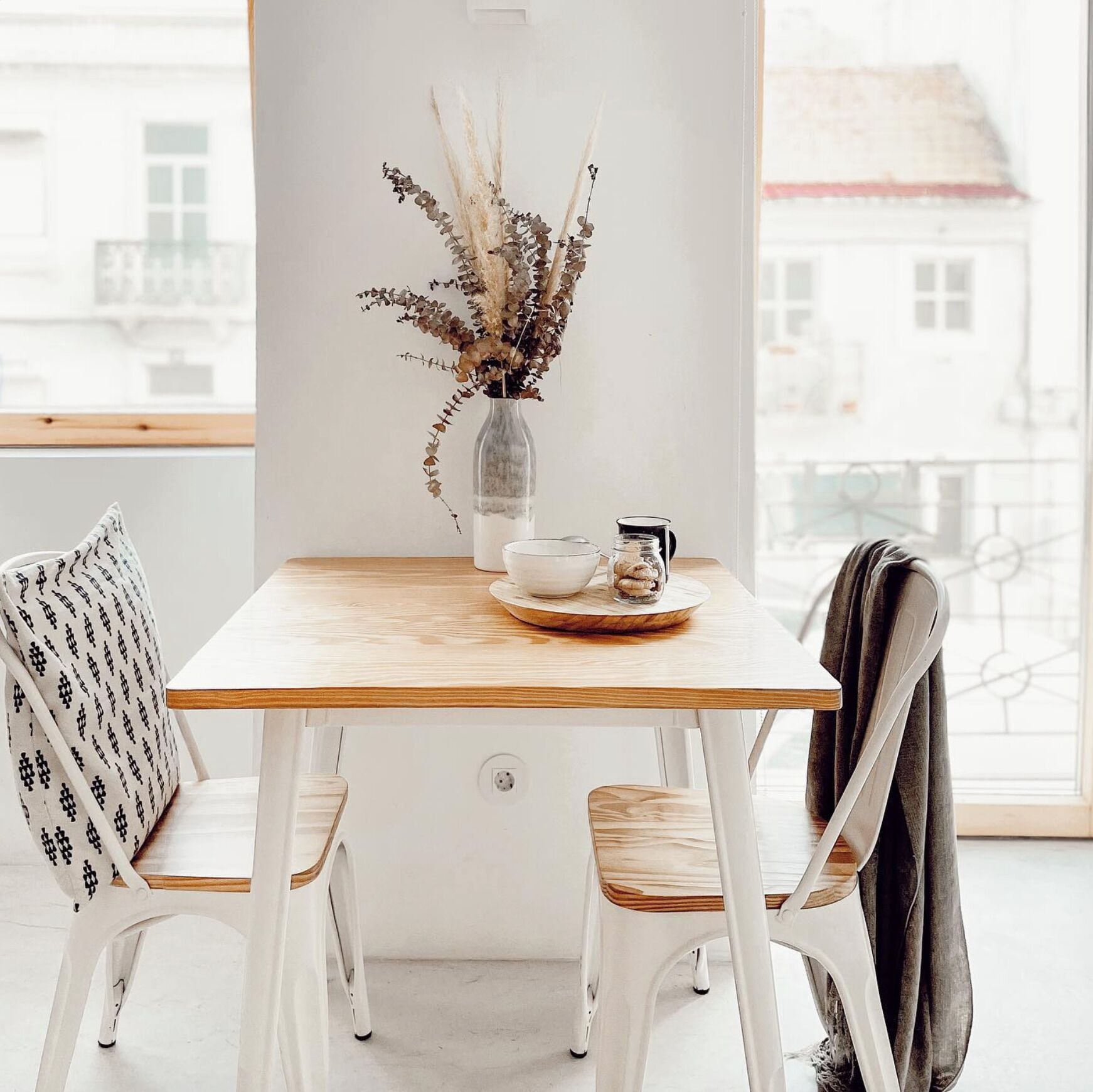 Un comedor minimalista con una mesa de madera, sillas blancas y un jarrón con plantas secas.