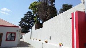 Une rue étroite bordée d’un immeuble blanc, avec une porte rouge et une petite fenêtre. Des plantes en pot sont disposées le long du mur, et un grand arbre se dresse à l’arrière-plan.