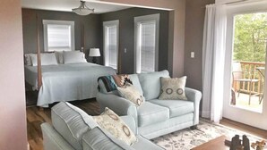 A bedroom with a bed, a sofa, and a chair. There is a window with blinds and a view of greenery outside.