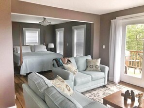 A bedroom with a bed, a sofa, and a chair. There is a window with blinds and a view of greenery outside.