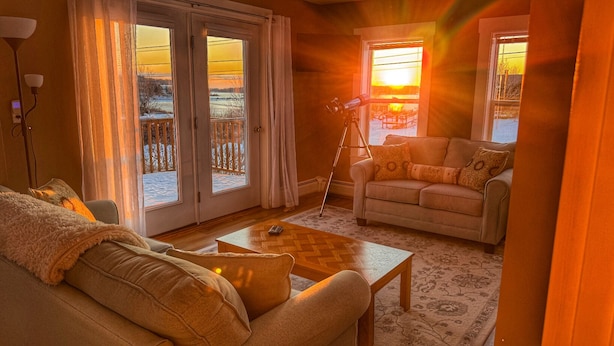 A cozy living room with a sofa, armchair, and coffee table. There is a telescope set up near a window with a view of a snowy landscape.