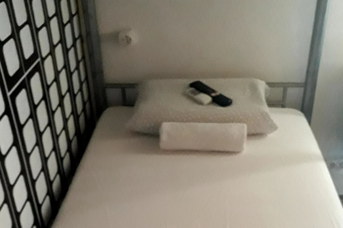Een enkel bed met een witte matras en een kussen, een klein nachtkastje met een lamp, en een raam met een metalen rooster.