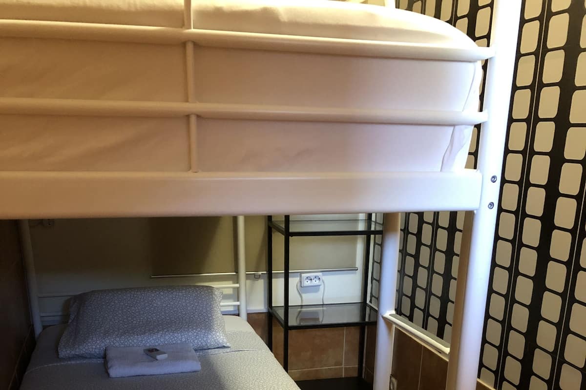 Een stapelbed met matras, kussens en dekbed. Er is een nachtkastje met een lamp en een stopcontact. De kamer heeft behang met een patroon en een betegelde vloer.