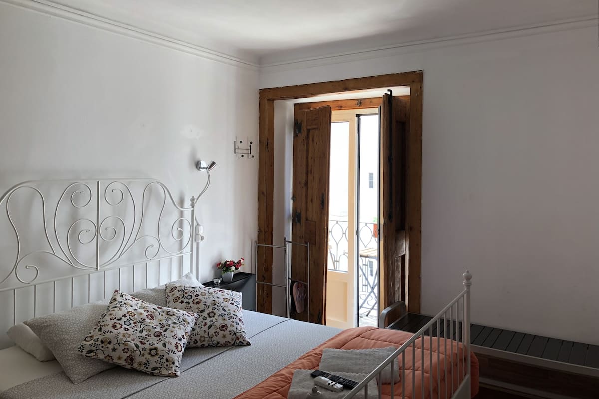 Een slaapkamer met een wit metalen bed, kussens met bloemenprint, een witte sprei en een houten deur naar een balkon.