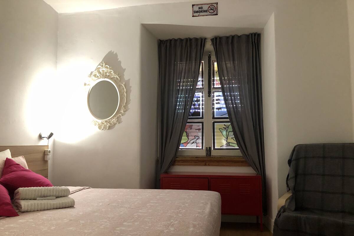 Een slaapkamer met een bed, een raam met gordijnen, een spiegel en een stoel.