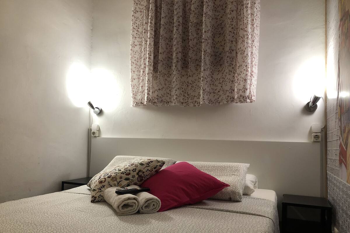 Een net opgemaakt bed met een sierkussen met bloemendessin, een opgerolde handdoek en een rood kussen. Op het bed ligt een afstandsbediening. De kamer heeft een witte muur met een bloemen gordijn en een wandlamp.