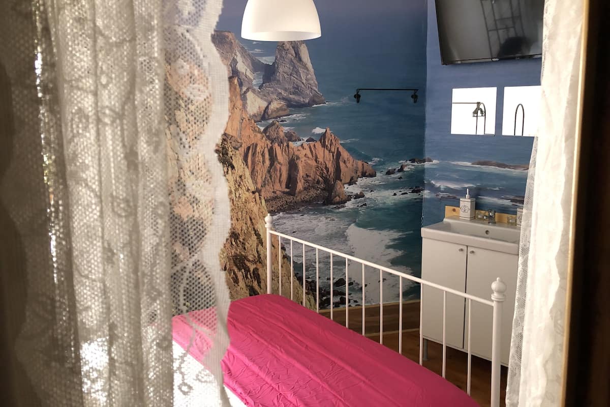 Een slaapkamer met een bed, een witte nachtkastje, een spiegel, een raam en een muur met een landschapsdecoratie.