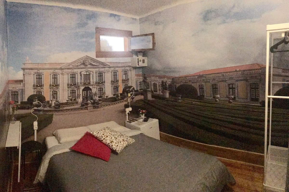 Een slaapkamer met een muurschildering van een klassiek gebouw, een bed met kussens, een nachtkastje en een hangende televisie.