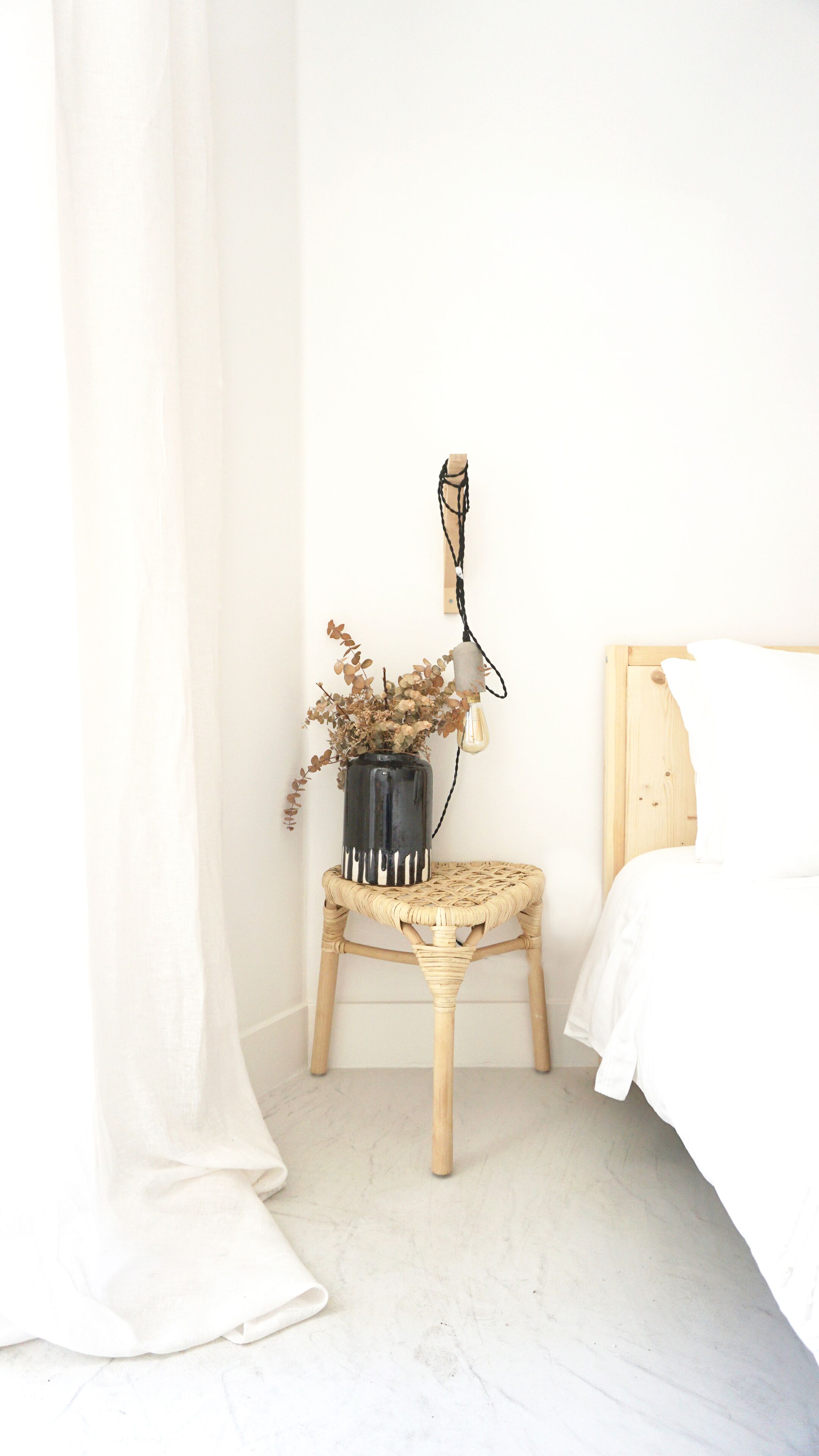 A bedroom with a bed, a small wooden stool with a vase of dried flowers, and a wall-mounted light fixture.