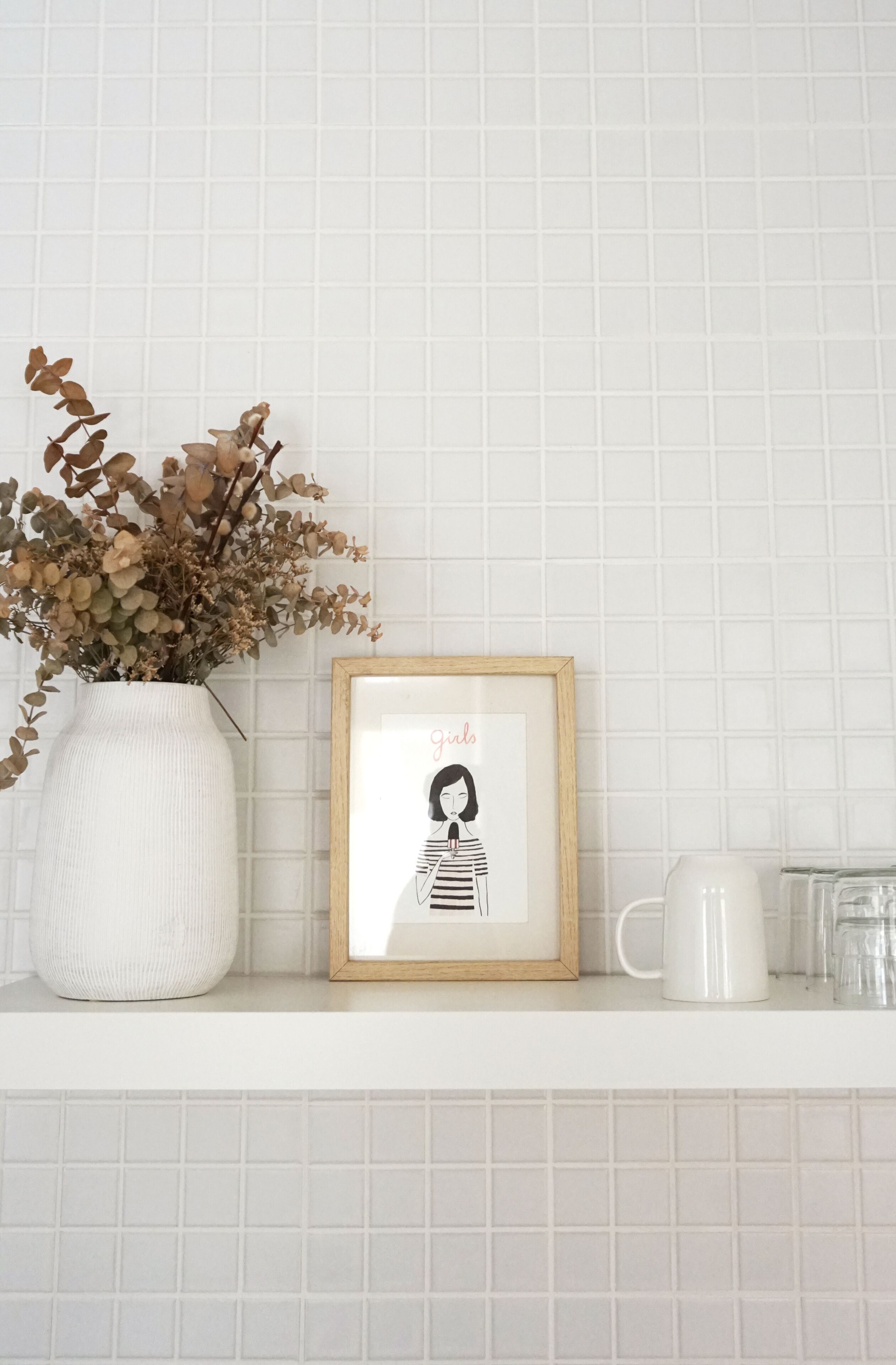 Un mur recouvert de carreaux blancs, orné d’un tableau encadré représentant une jeune fille et d’un vase blanc contenant des fleurs séchées.