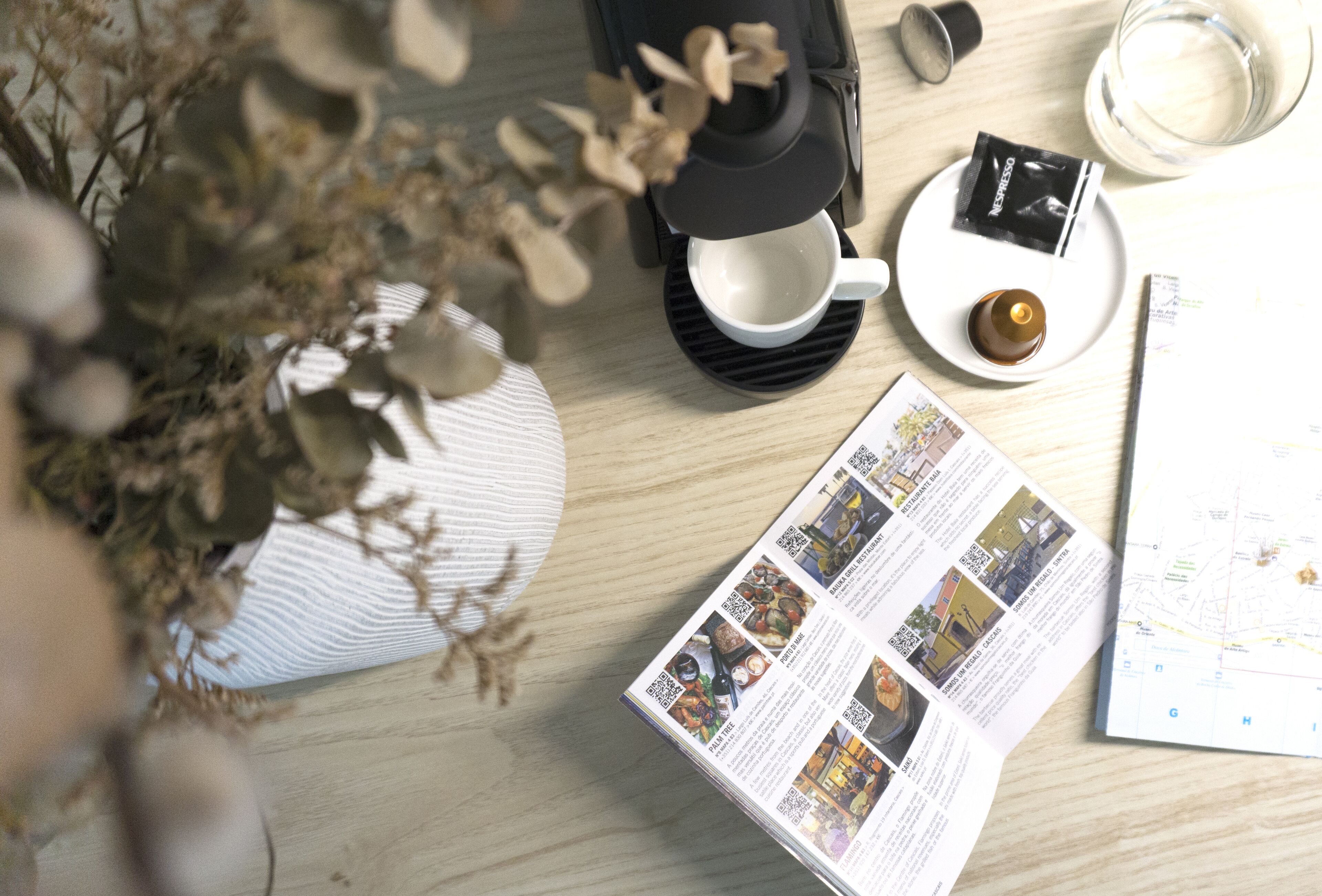 A table with a coffee machine, a cup, a plate with a packet, a candle, a magazine, and a map.