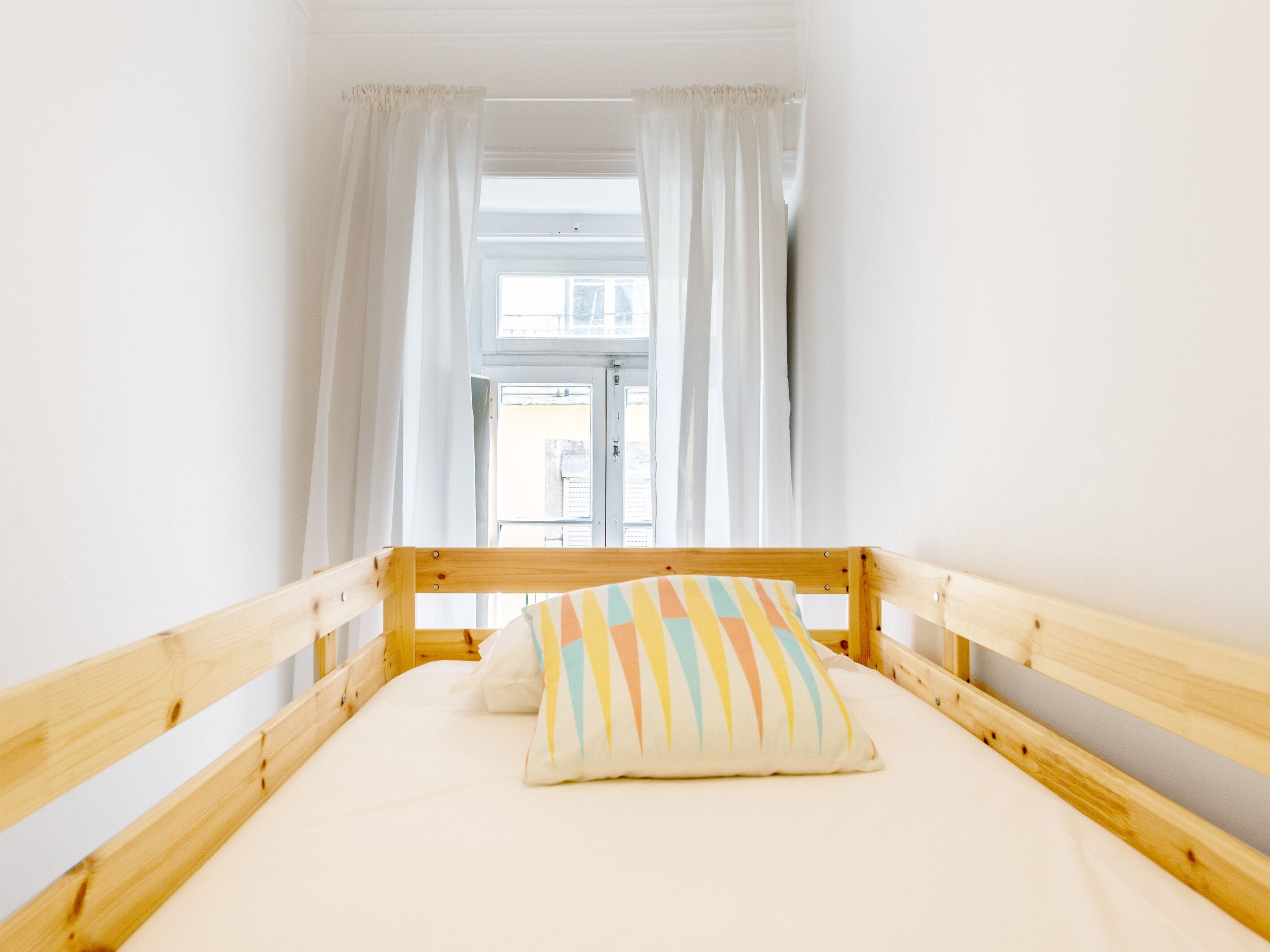 Una habitación pequeña con cama individual, estructura de madera, ropa de cama blanca y almohada colorida.