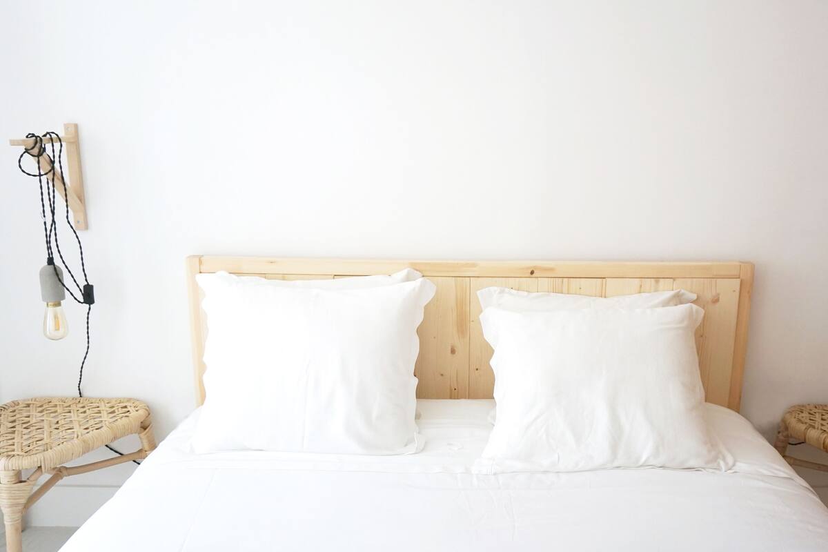 Una cama con cabecera de madera, dos almohadas blancas y dos mesitas de noche de mimbre con lámparas.