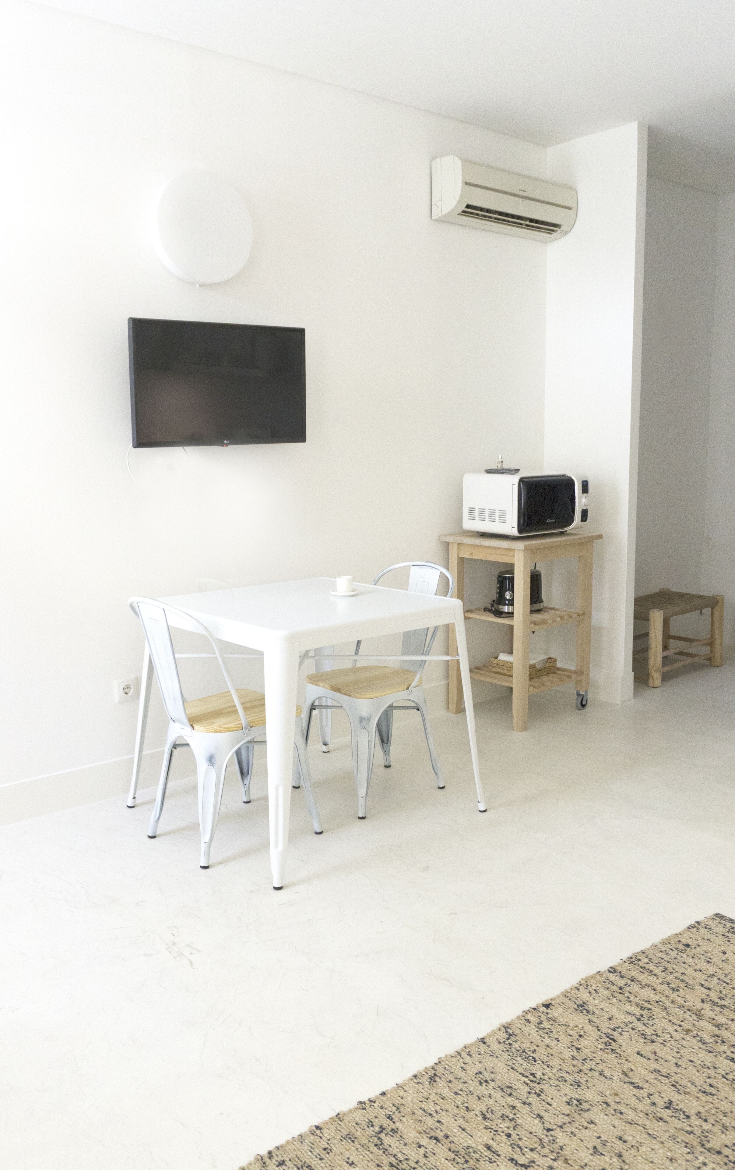 Ein minimalistisch eingerichteter Raum mit einem weißen Tisch, zwei Stühlen, einem kleinen Holztisch mit einer Mikrowelle und einer weißen Klimaanlage an der Wand.